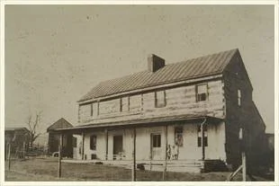 Old two-story wooden house with a porch in the foreground, likely from the early 20th century.