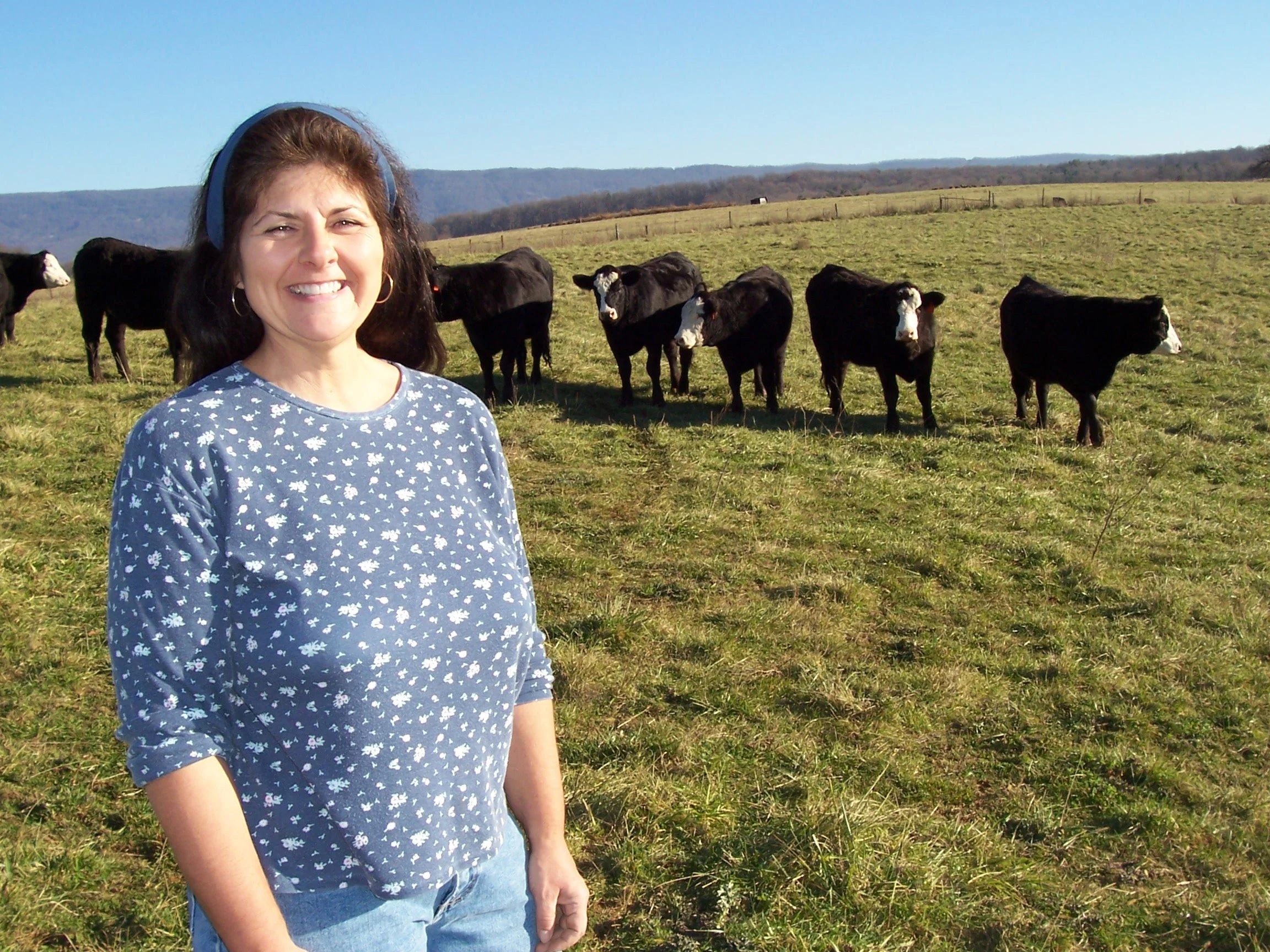 Woman smiling outdoors in a field with cows grazing in the background under a clear blue sky.