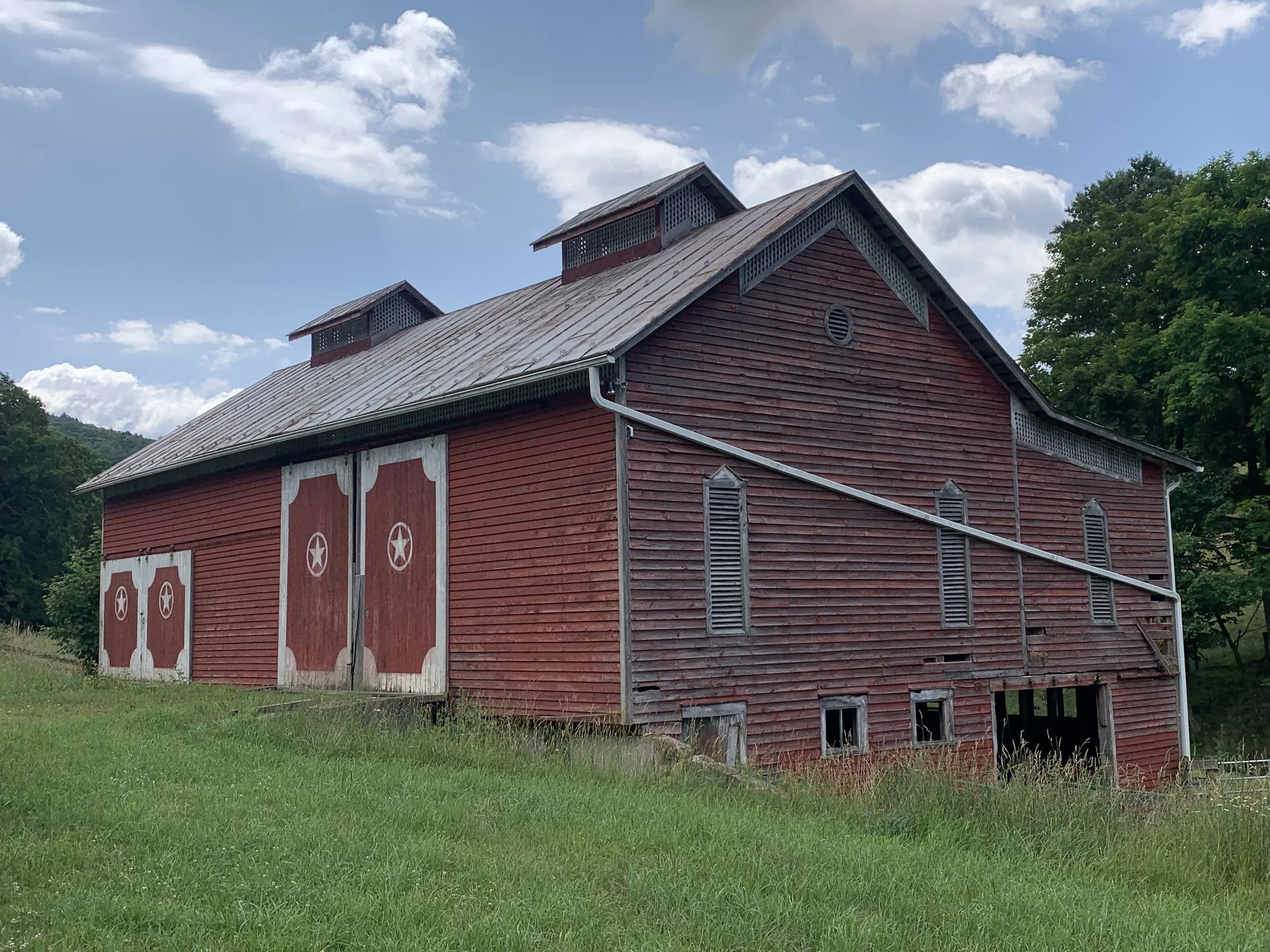 A red barn with white accents and double doors, with three small windows on the side and two attic vents, set in a grassy landscape under a partly cloudy sky.