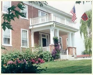 A brick house with a front porch and white railings, decorated with American flags and a Confederate flag, surrounded by well-maintained bushes and flowers.