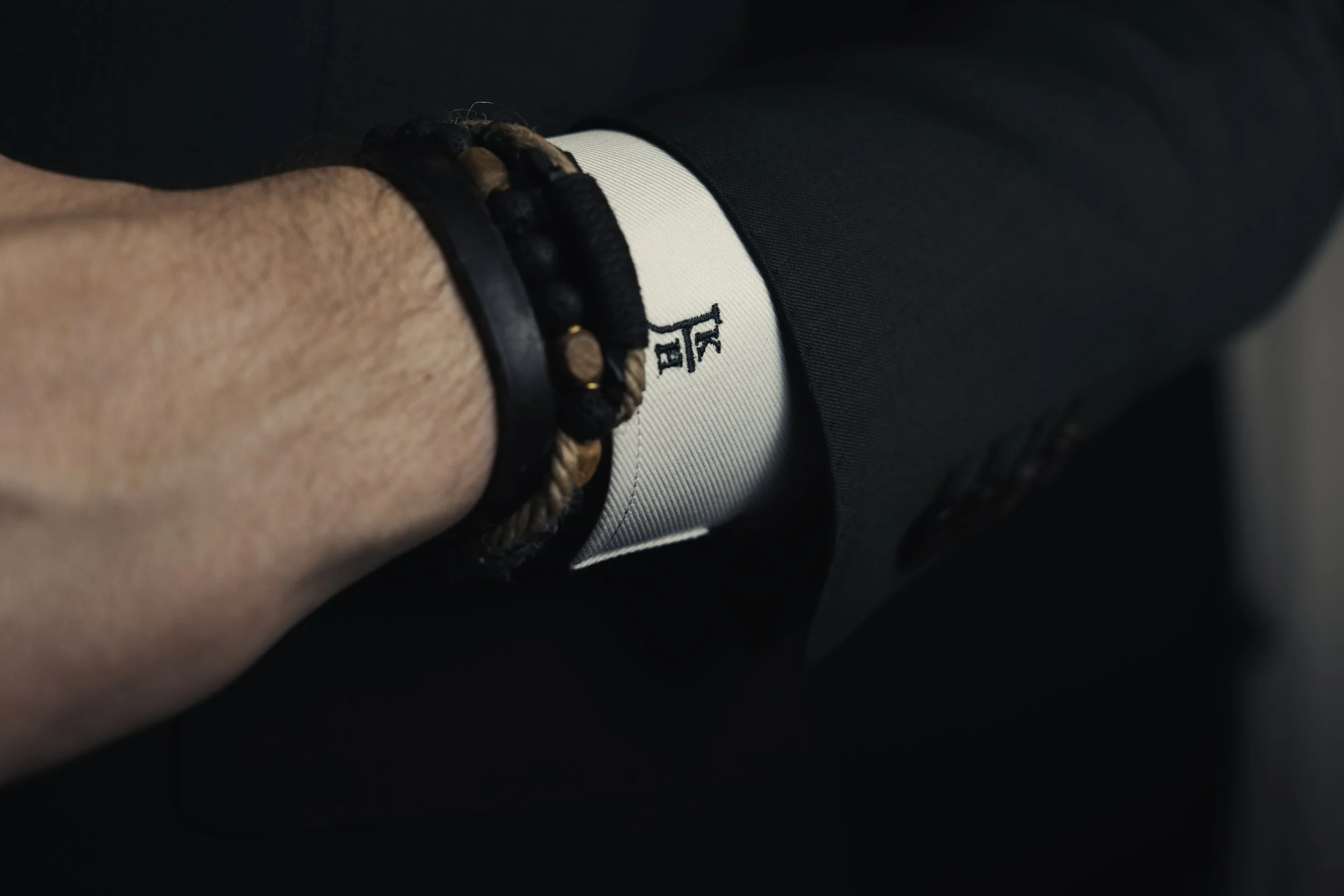Close-up of a person's wrist wearing black leather and woven beaded bracelets, with a glimpse of white clothing and dark green suit fabric.