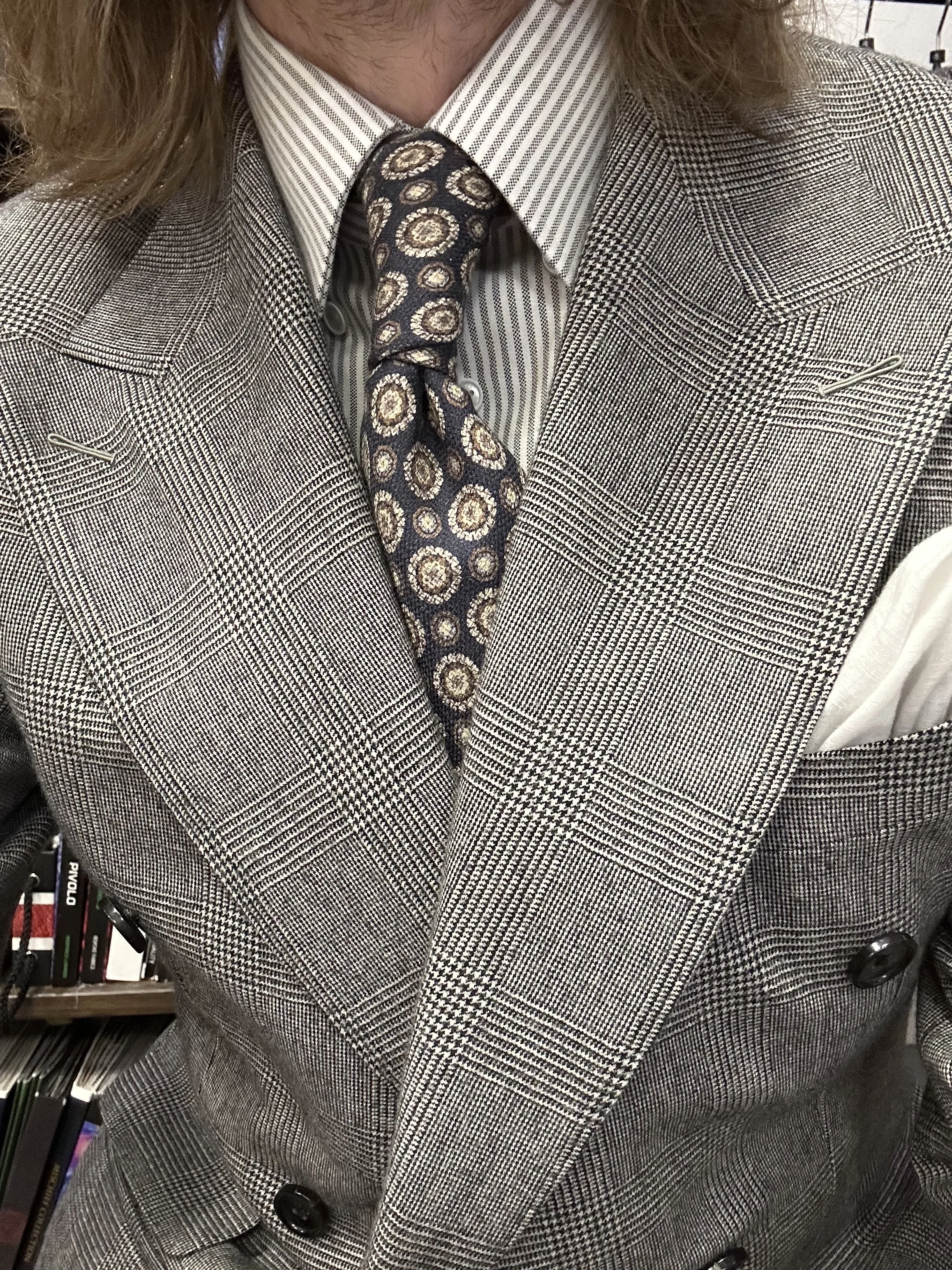 Close-up of a person wearing a glen plaid suit, a striped shirt, and a patterned tie, with books and office supplies in the background.