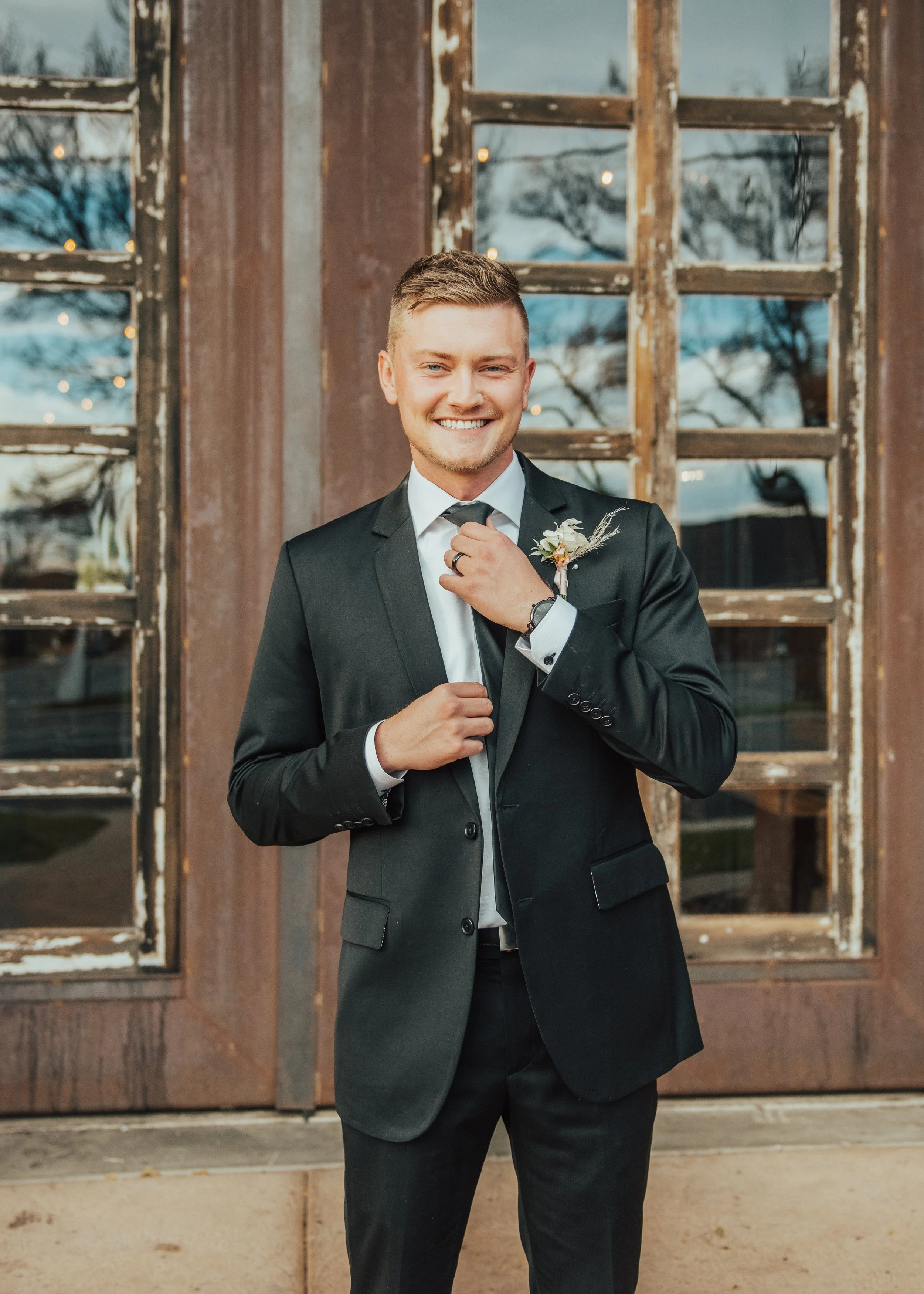 A man in a formal black suit and white shirt smiling and adjusting his tie, standing in front of a weathered wooden window with a winter landscape outside.