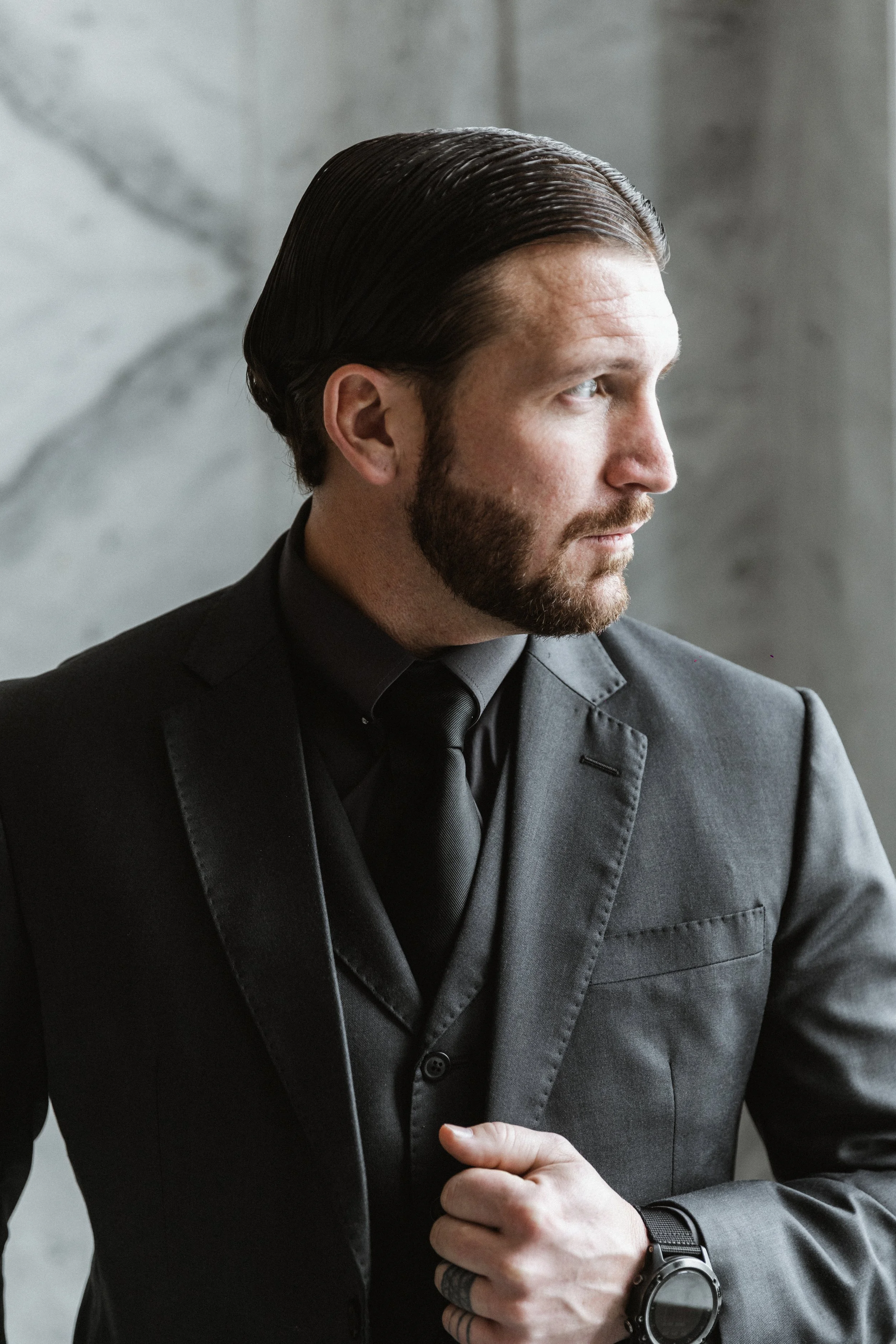 Side profile of a man with slicked-back hair and a beard, wearing a dark grey suit, dark grey shirt, dark grey tie, and a black wristwatch, standing in front of a grey marble background.