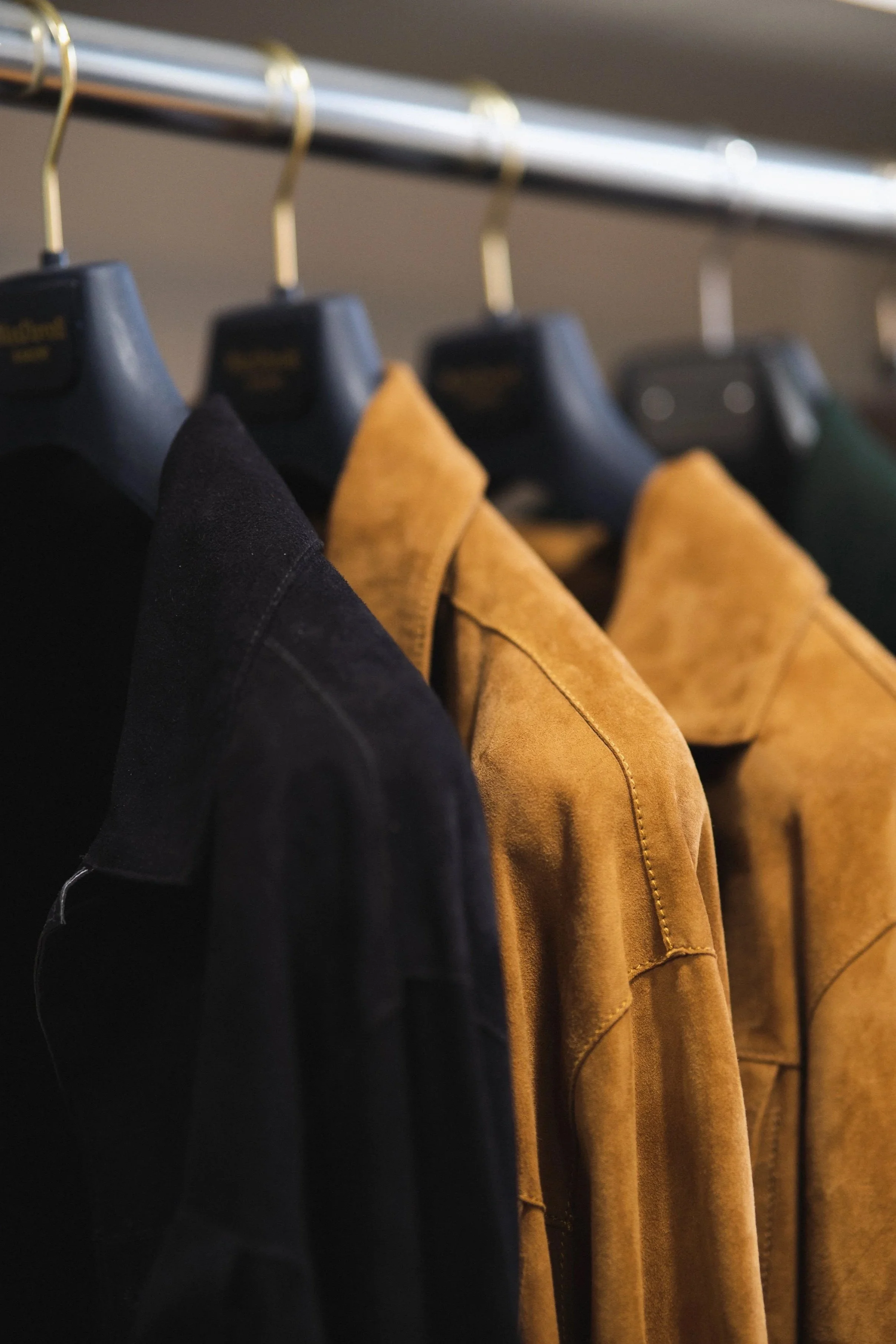 Close-up of hanging black and tan jackets on black and gold hangers, on a metal clothing rod, with a blurred background.
