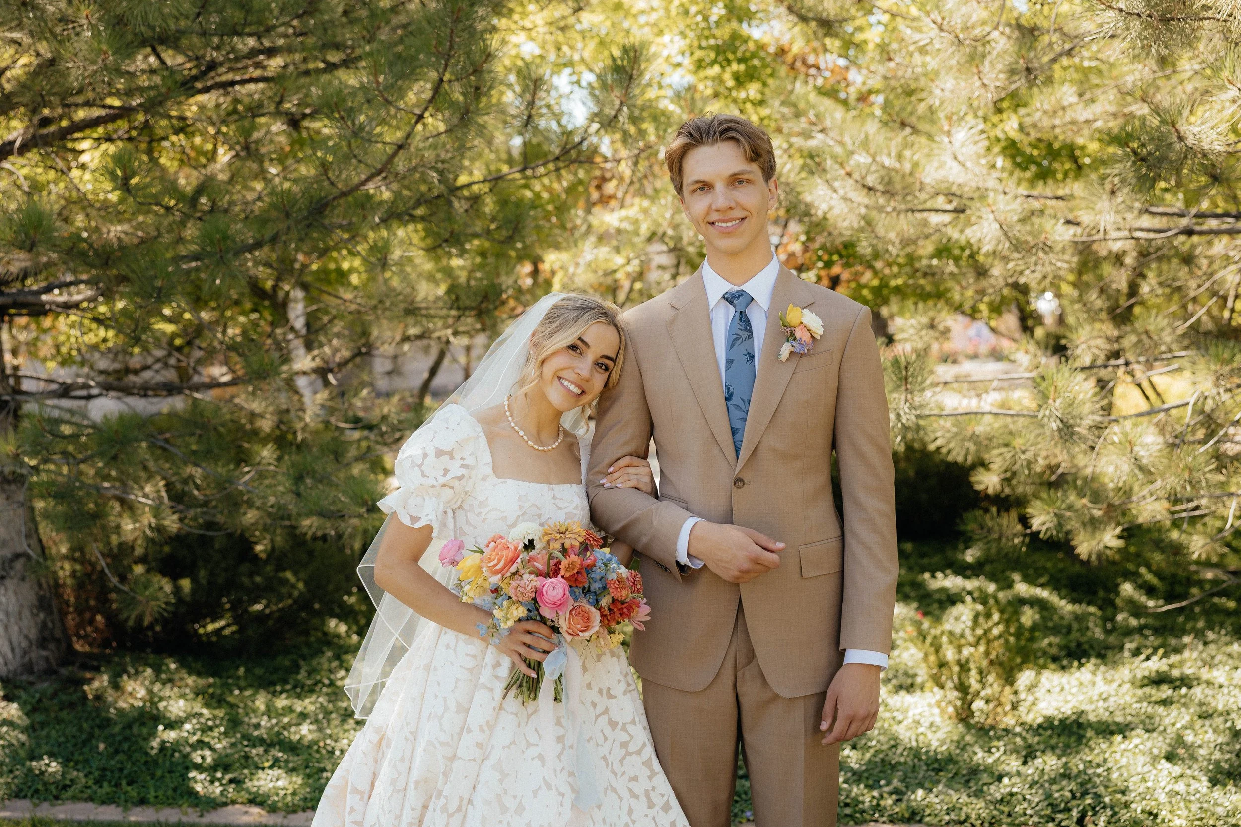 A bride and groom smiling outdoors among trees, with the bride holding a colorful bouquet and the groom wearing a tan suit and blue tie.
