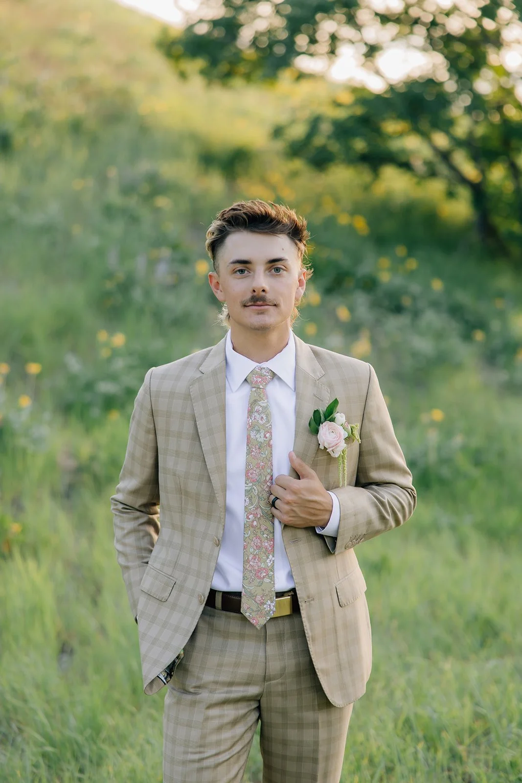 A young man dressed in a beige plaid suit with a floral tie, standing outdoors in a grassy area with trees in the background. He has a floral boutonniere on his lapel and is touching his chest with one hand.