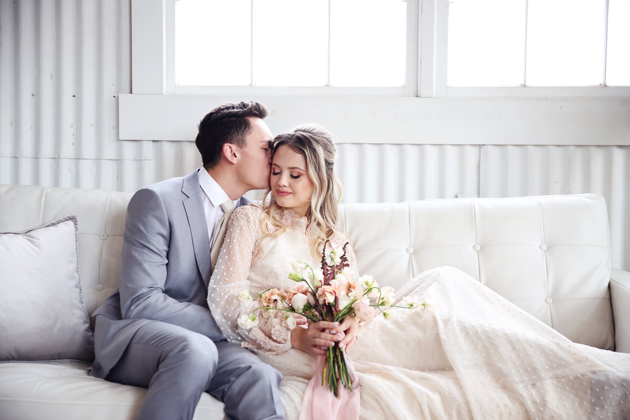 A bride and groom sitting on a white couch in a bright, minimalistic room. The groom is kissing the bride on the forehead, and she is holding a bouquet of flowers. The room has large windows and white walls.