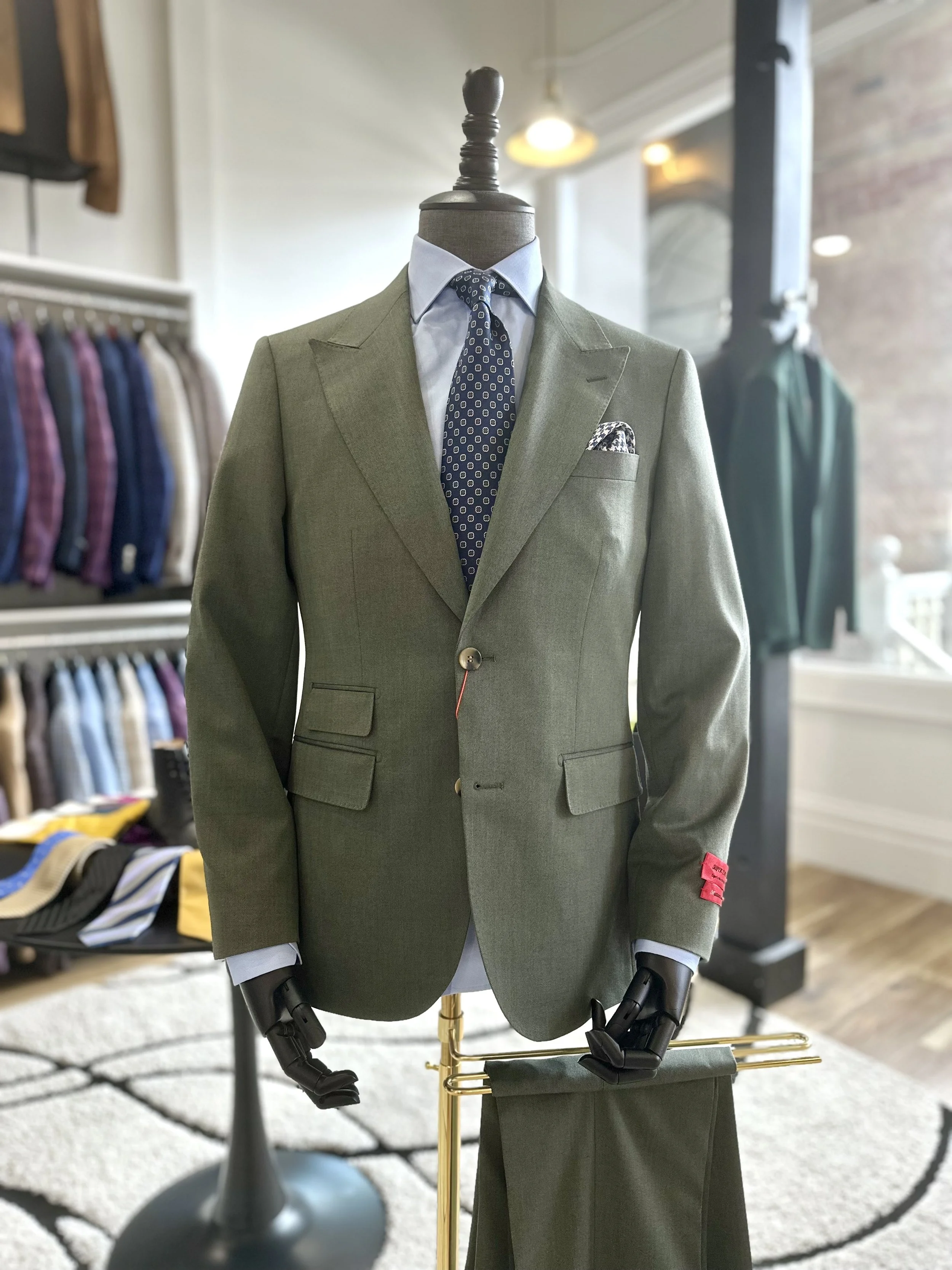 A mannequin dressed in a green suit with a light blue shirt, a patterned blue tie, and a pocket square, displayed in a clothing store.