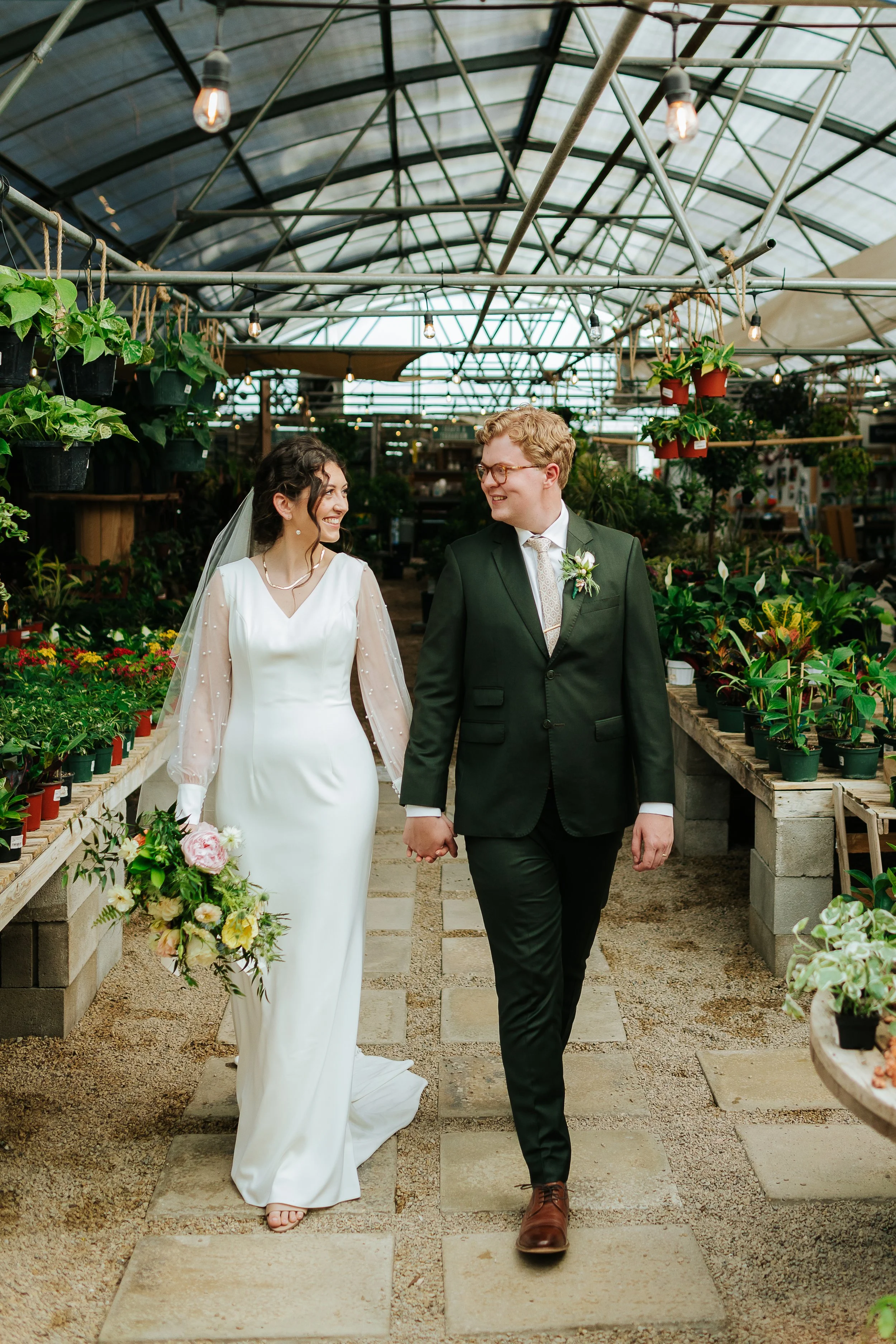 A bride and groom walking hand in hand through a greenhouse decorated with plants and flowers, smiling at each other.