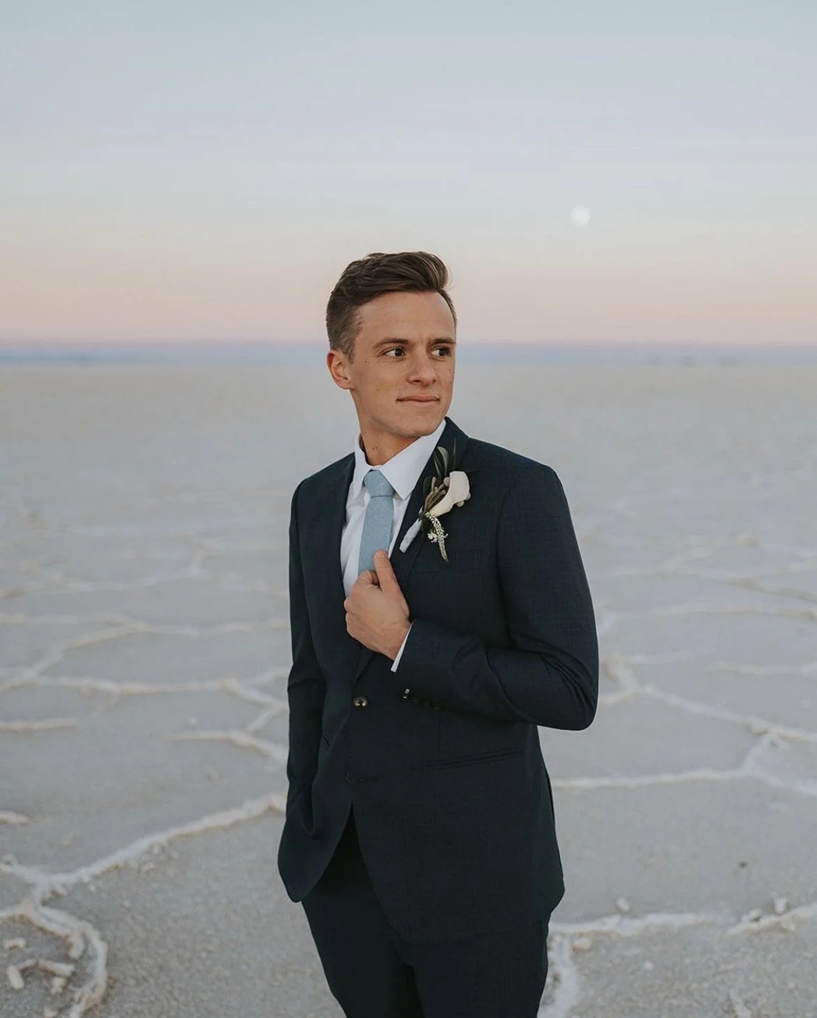 A man in a dark suit with a light blue tie and white shirt standing outdoors on a salt flat during sunset, holding a flower lapel pin on his suit.