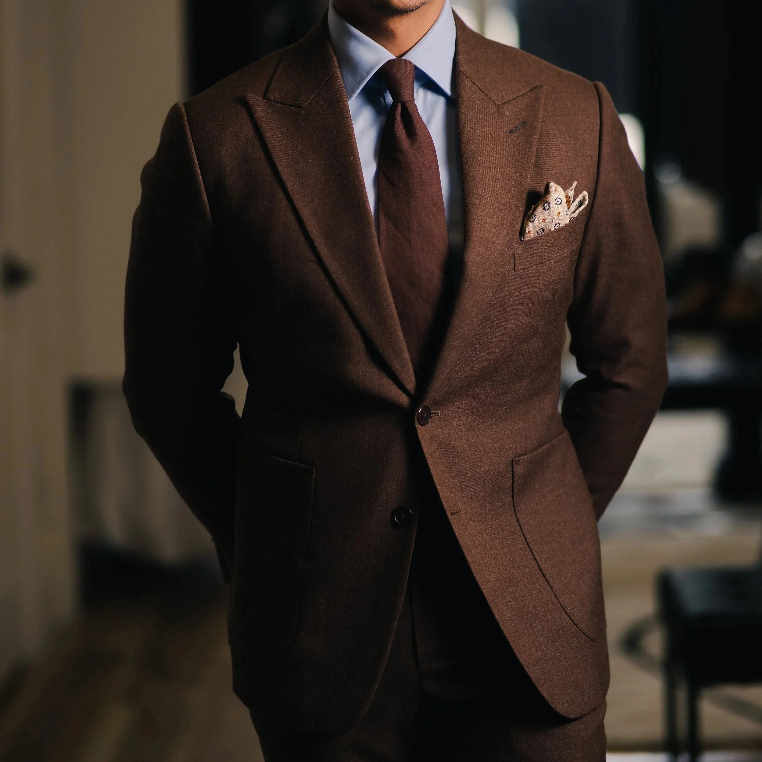 Close-up of a man wearing a formal brown/green flannel suit with a light blue shirt, brown tie, and a patterned pocket square. The image emphasizes the man's attire, with the focus on the suit and accessories.