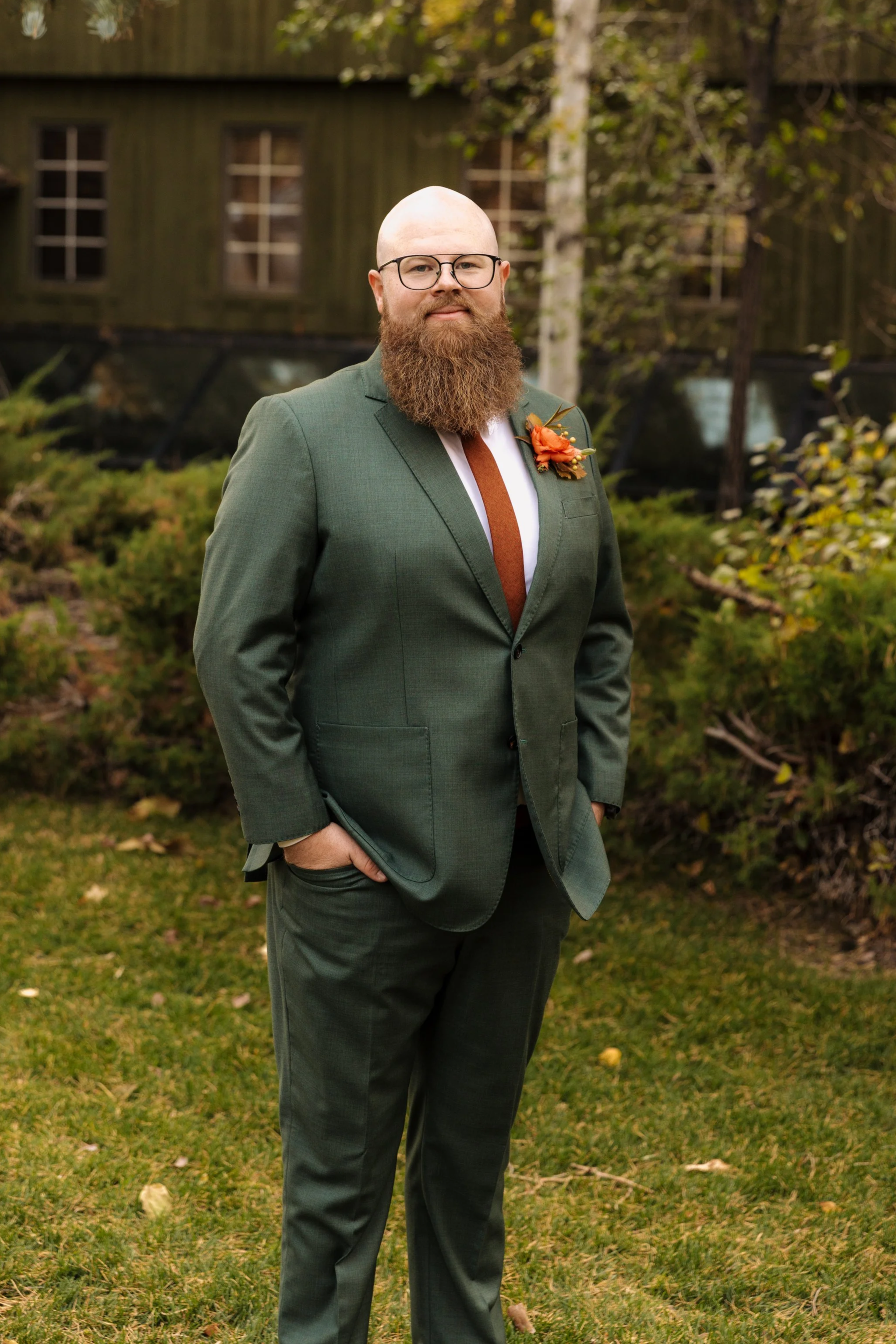 A man with glasses, a bald head, and a full beard wearing a green suit with a white shirt and a brown tie, standing outdoors in front of a rustic building with window openings and trees with autumn leaves.