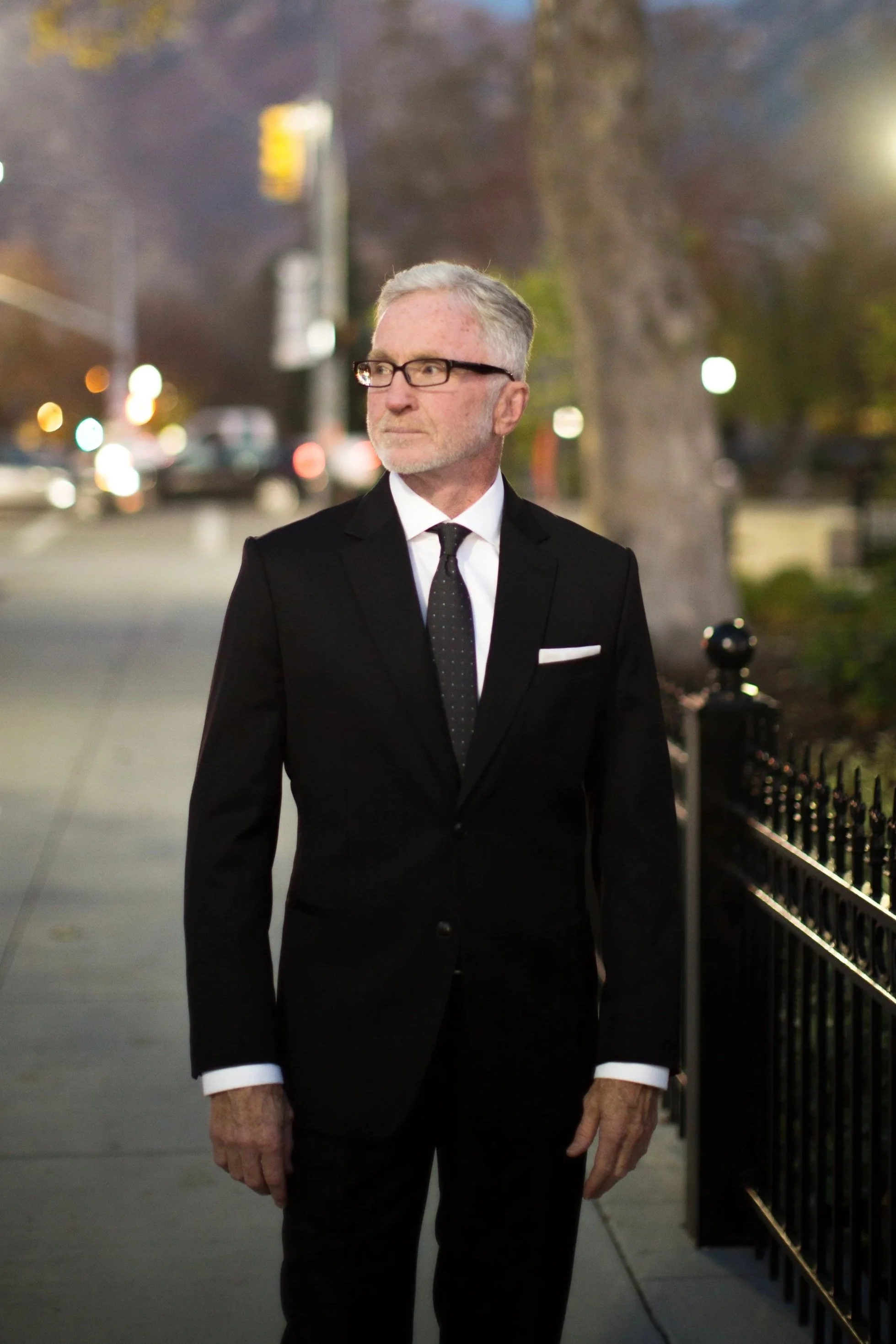 An elderly man with gray hair and glasses dressed in a black suit, white shirt, and black tie, standing on a sidewalk at night near a black metal fence with blurred city lights in the background.