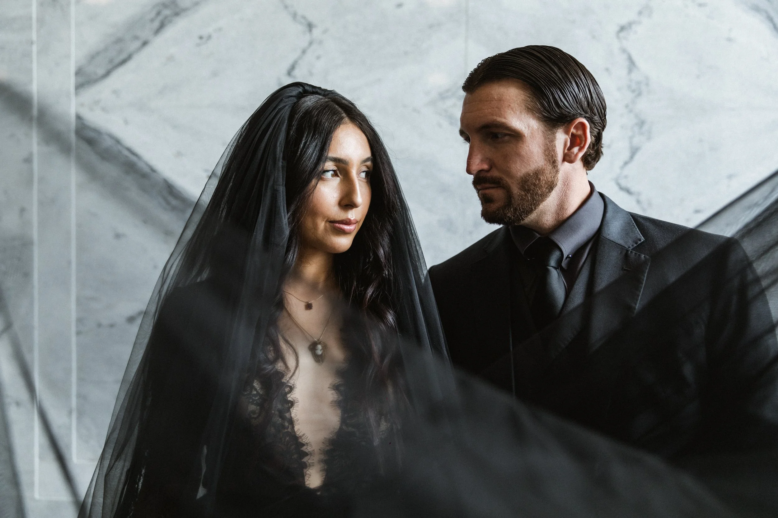 A man and a woman face each other closely in front of a marble wall. The woman has long dark hair, a black veil, and wears a lace top with a necklace. The man has slicked-back hair and a beard, dressed in a dark suit with a shirt and tie.