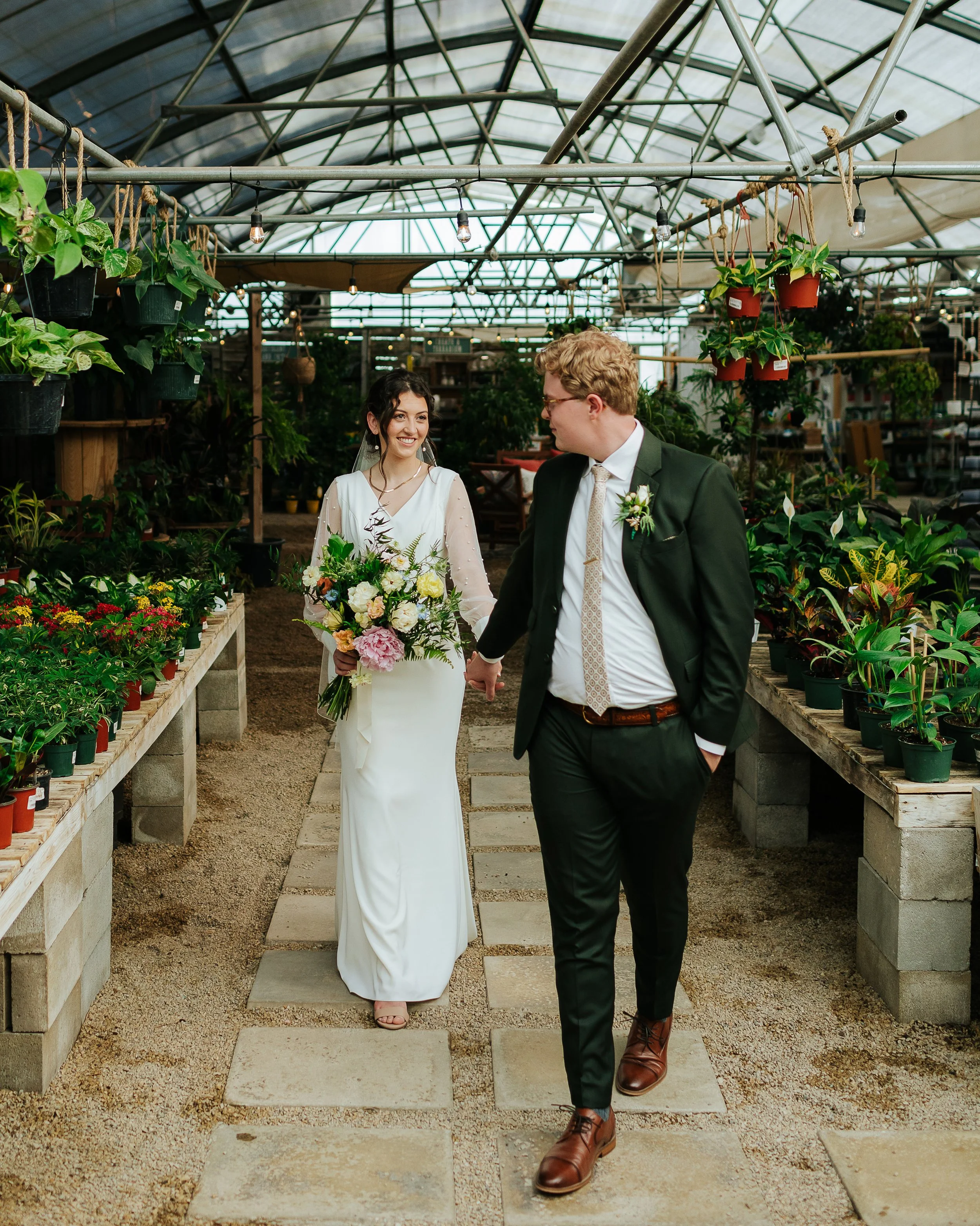 A bride and groom walking hand in hand through a greenhouse filled with lush green plants and flowers, with the bride carrying a bouquet, and both dressed in formal wedding attire.