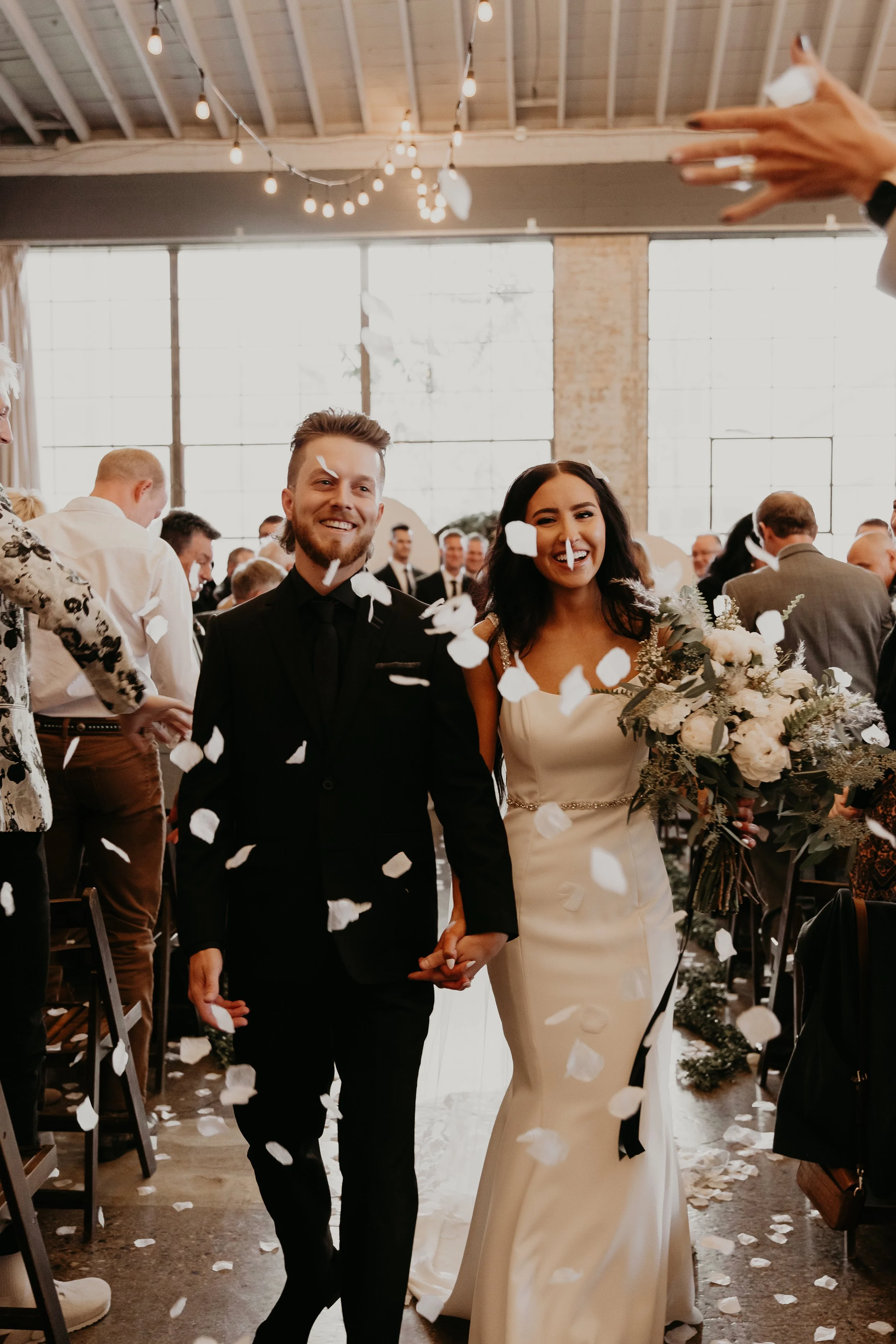 A bride and groom holding hands and smiling at their wedding ceremony, with confetti falling around them in a bright, industrial-style venue.