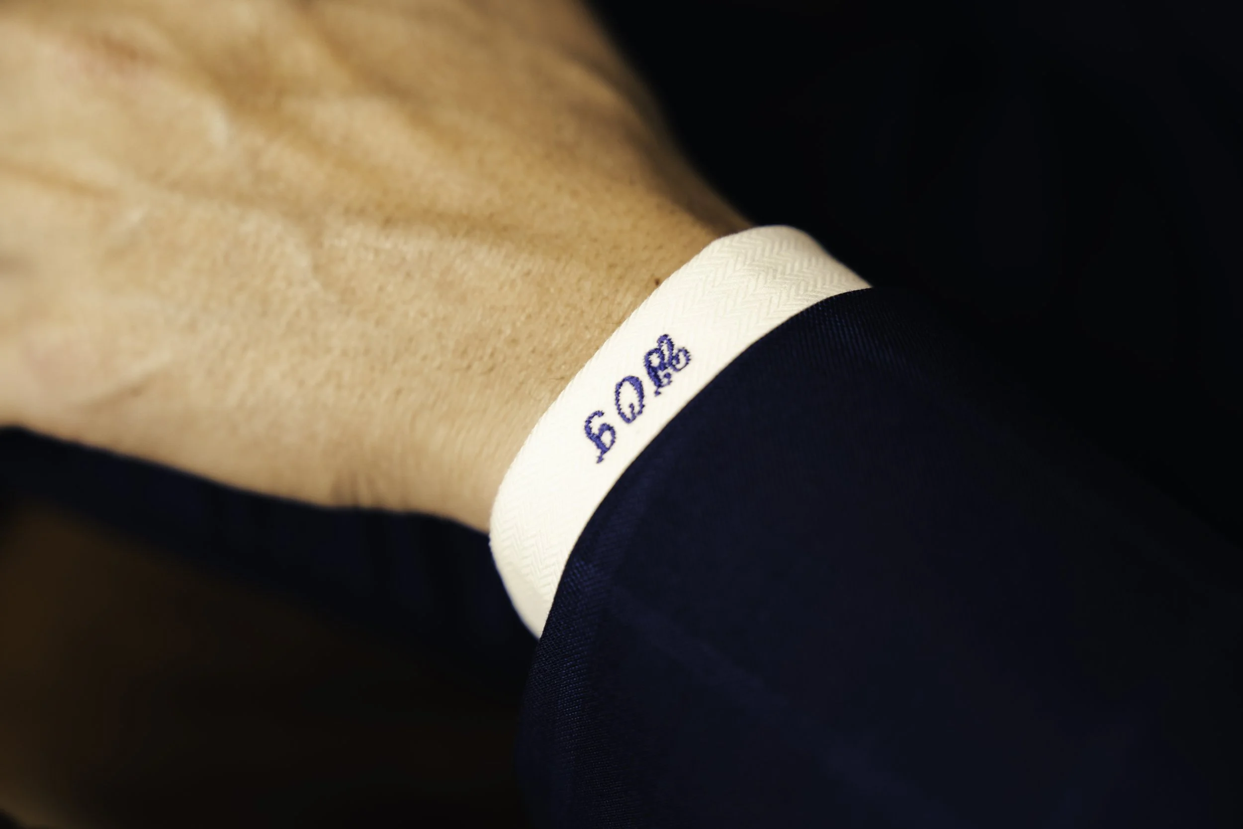 Close-up of a person's wrist with a white shirt cuff and dark blue suit, showing a logo embroidered on the cuff.