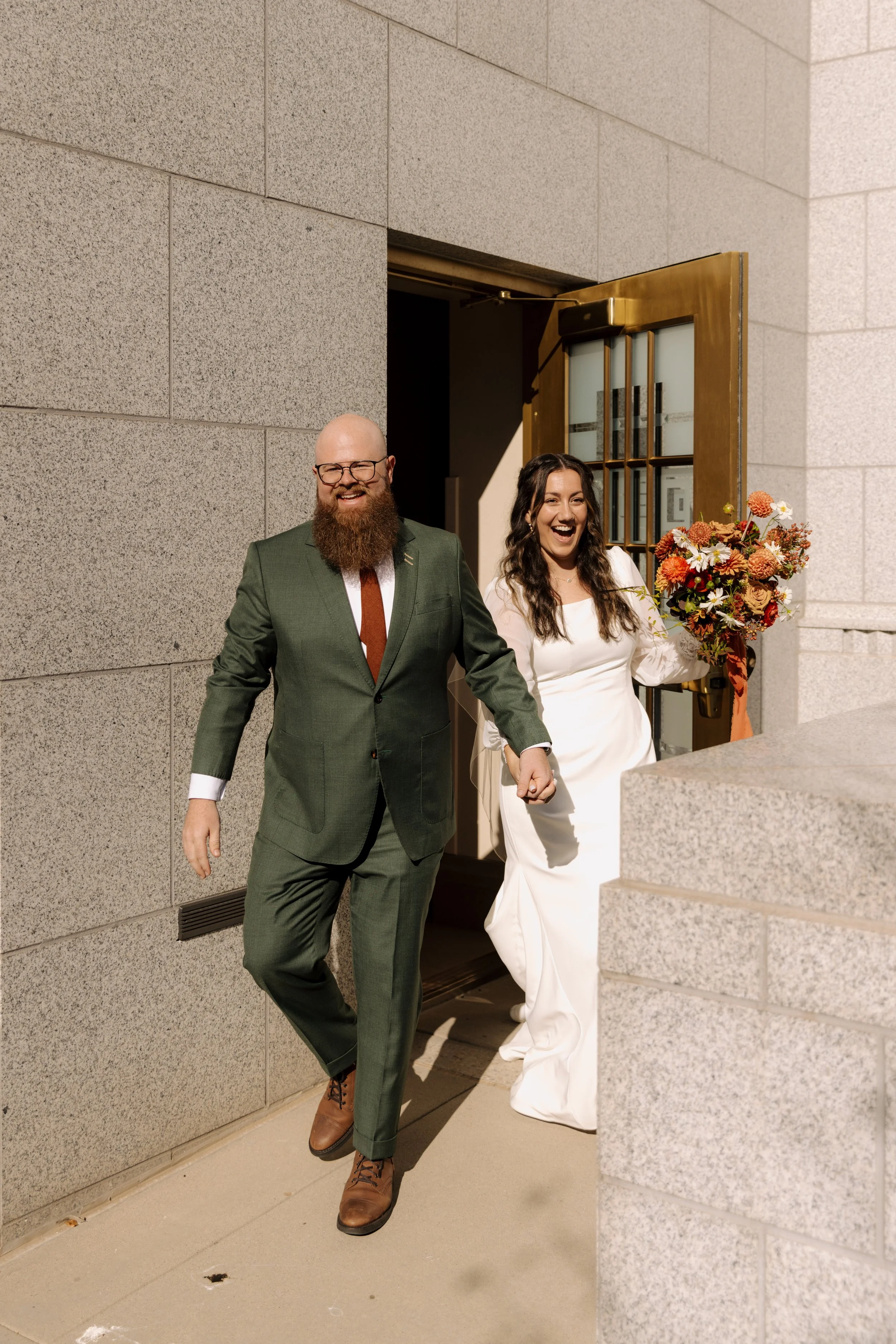 A joyful man and woman holding hands exit a building through a large door, the woman holding a bouquet of flowers, on a sunny day.
