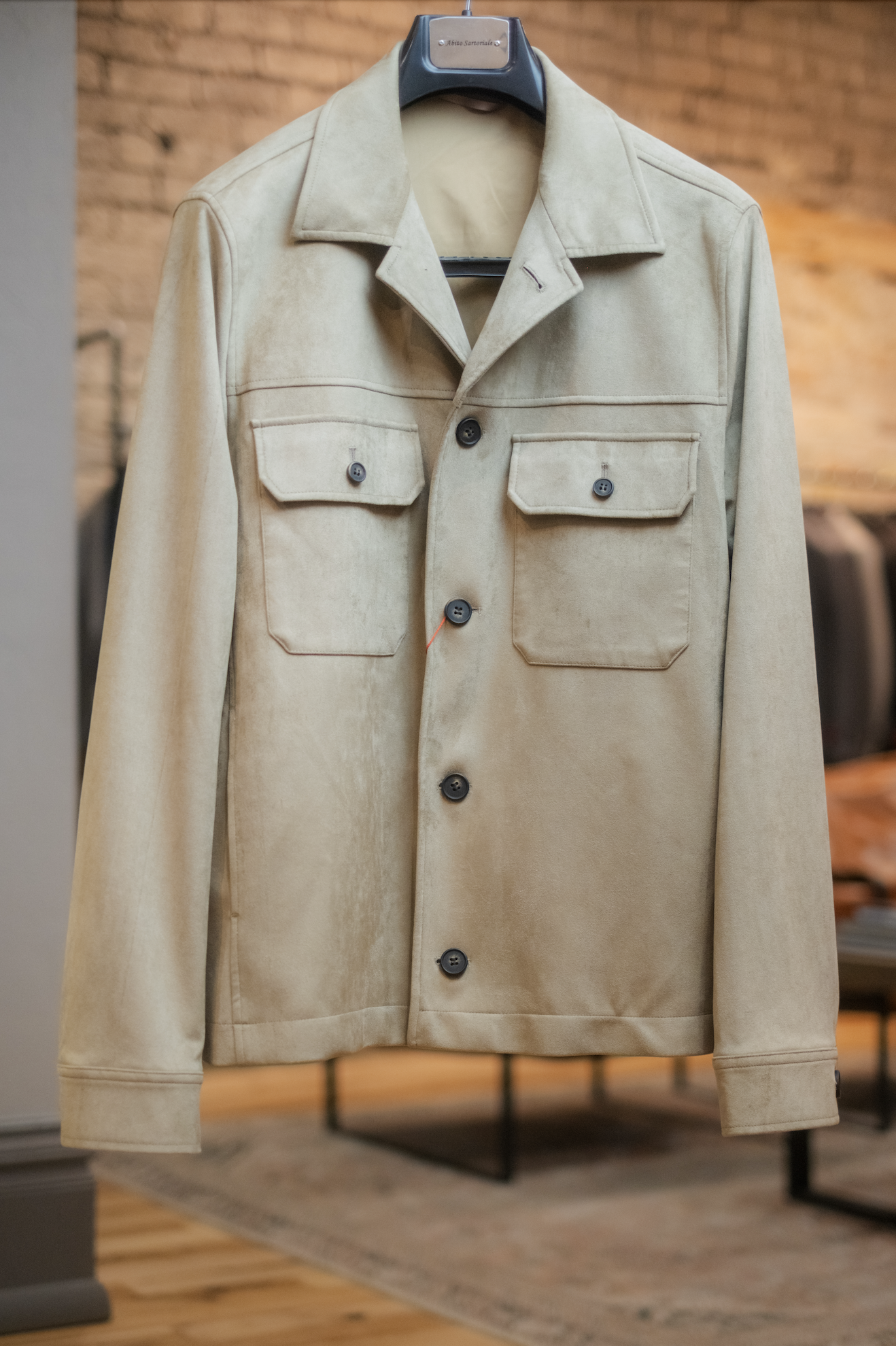 Beige jacket hanging on a black hanger against a brick wall background.