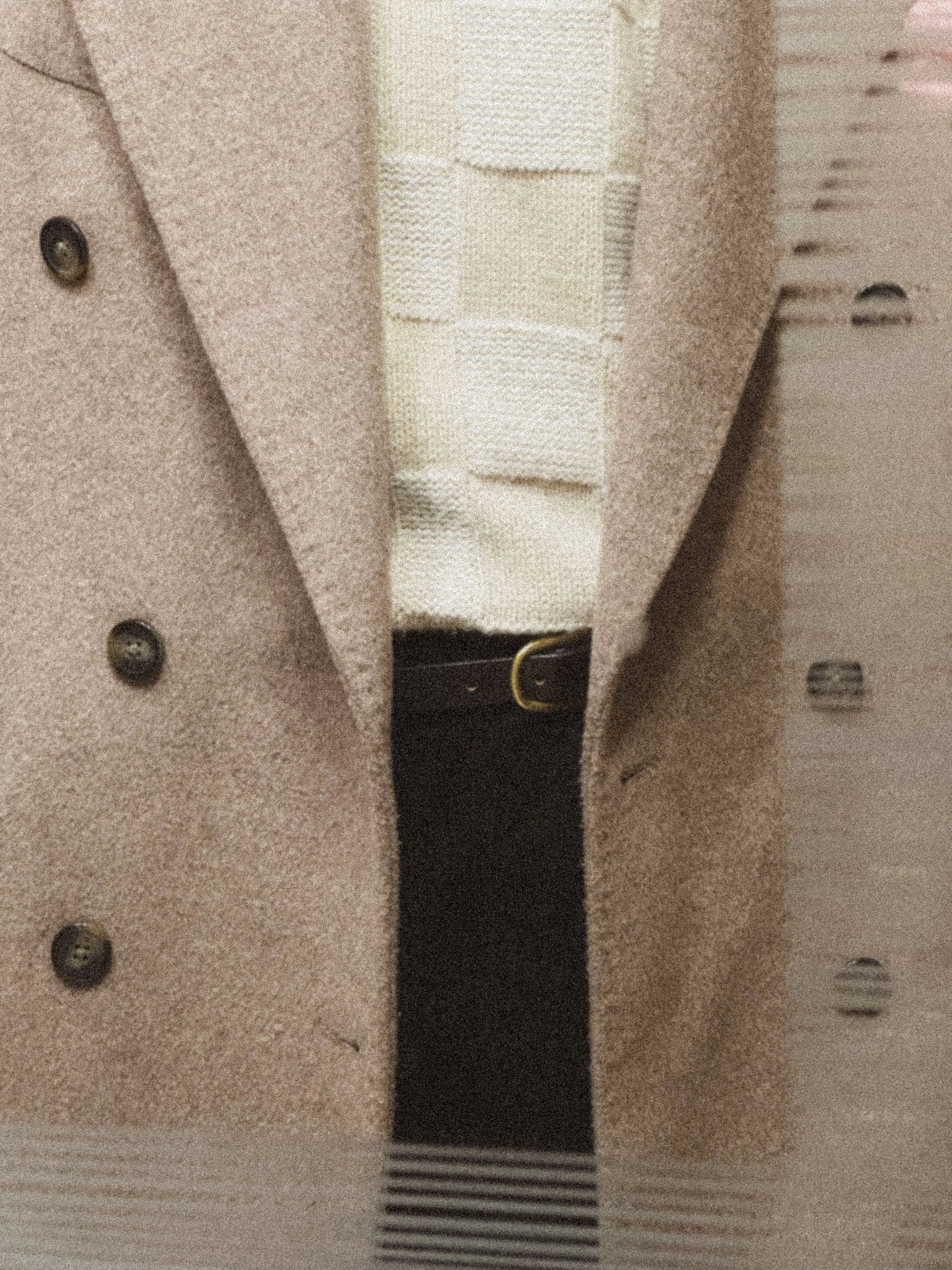Close-up of a beige overcoat with dark buttons, a cream sweater with a textured pattern, and a black belt with a gold buckle.