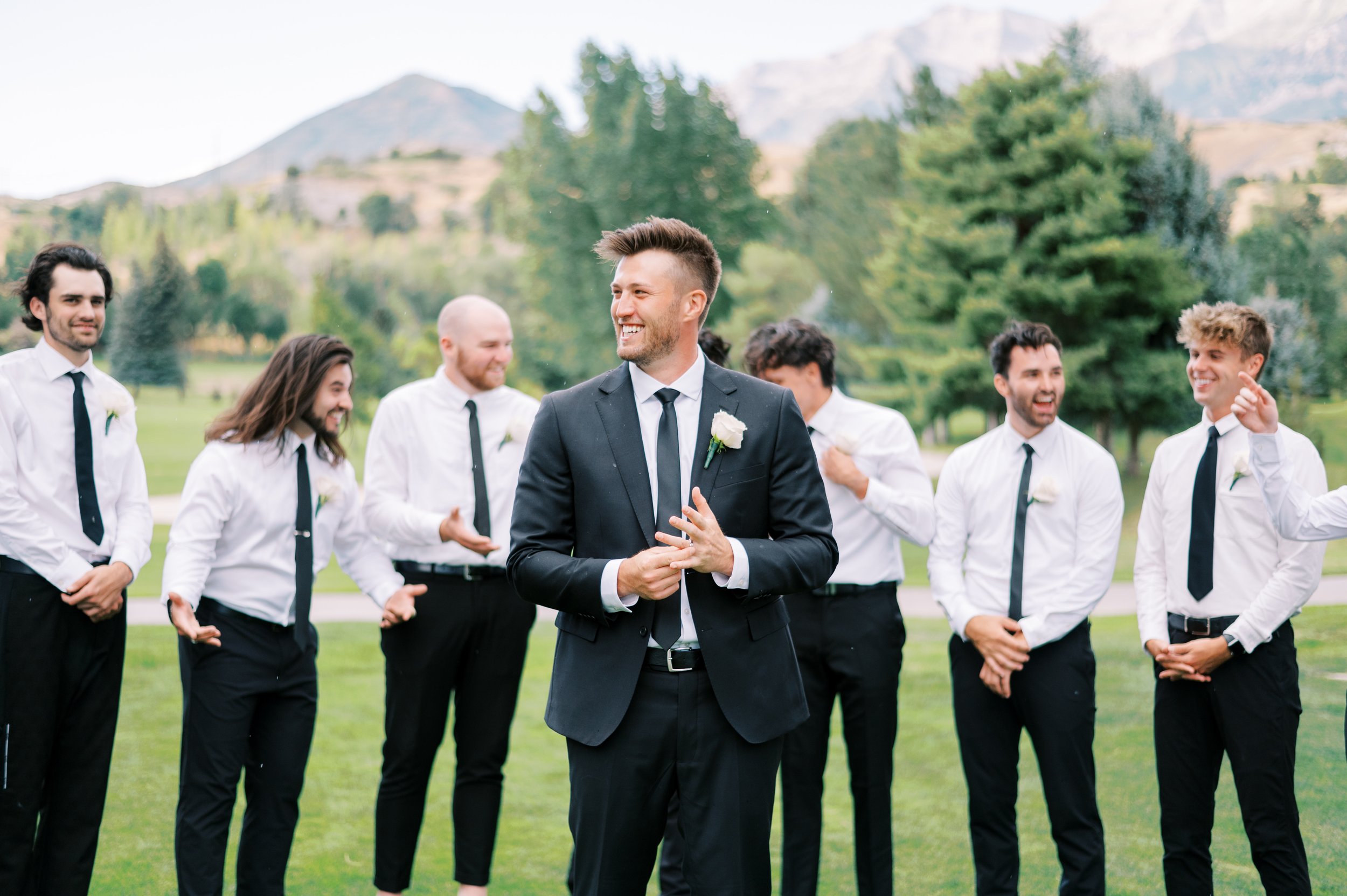 A groom in a suit smiling and holding hands together, surrounded by groomsmen in white shirts and black ties, outdoors with mountains and trees in the background.