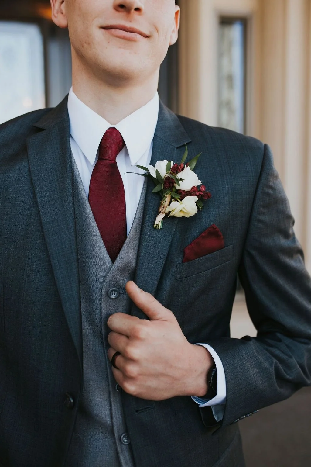 Close-up of a groom wearing a dark gray suit, white shirt, and maroon tie, adjusting his suit. He has a floral boutonniere pinned to his lapel and a matching maroon pocket square.