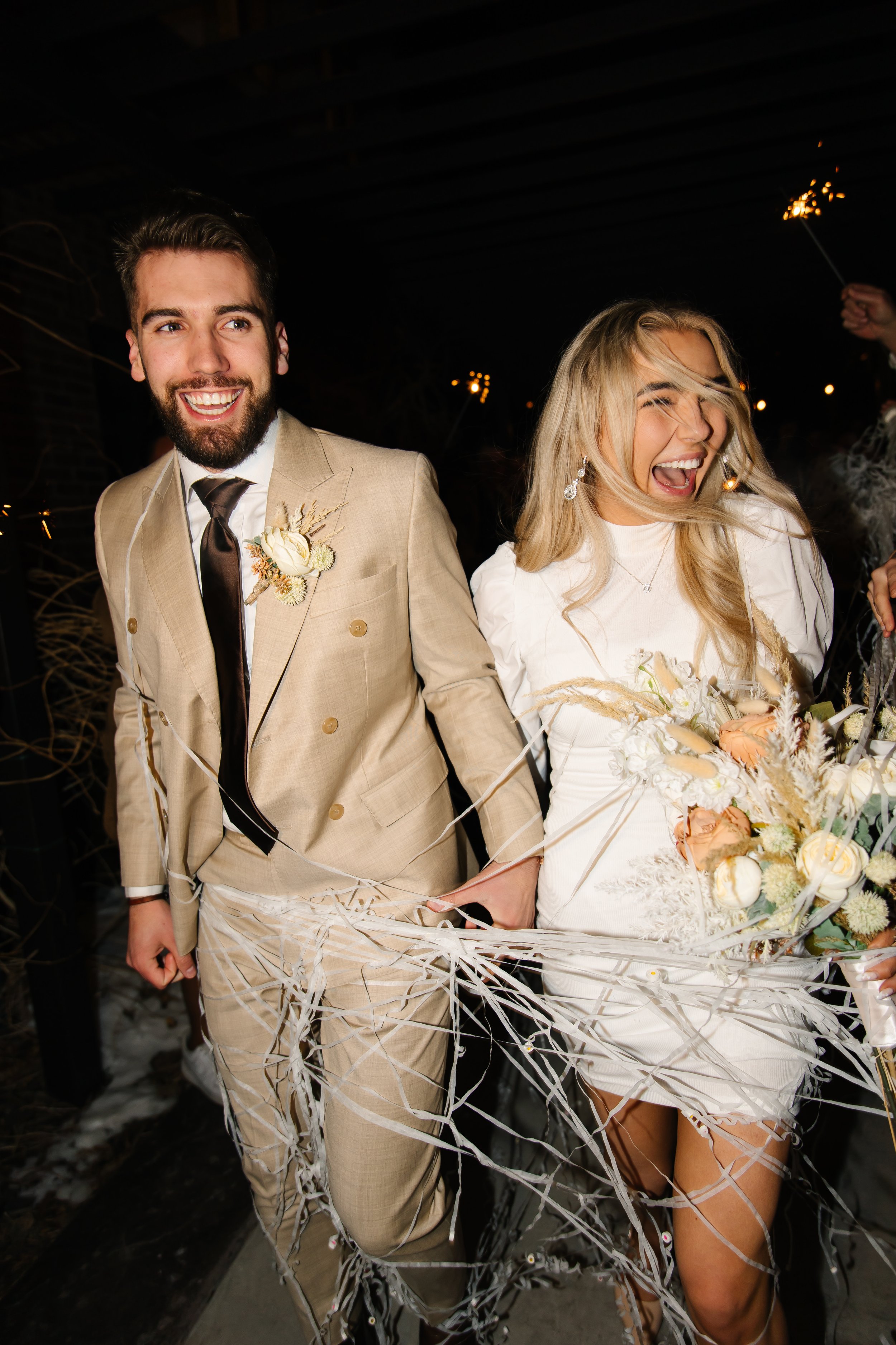 A joyful couple holding hands, dressed in wedding attire, walking through paper streamers at a celebration night event, with sparklers in the background.