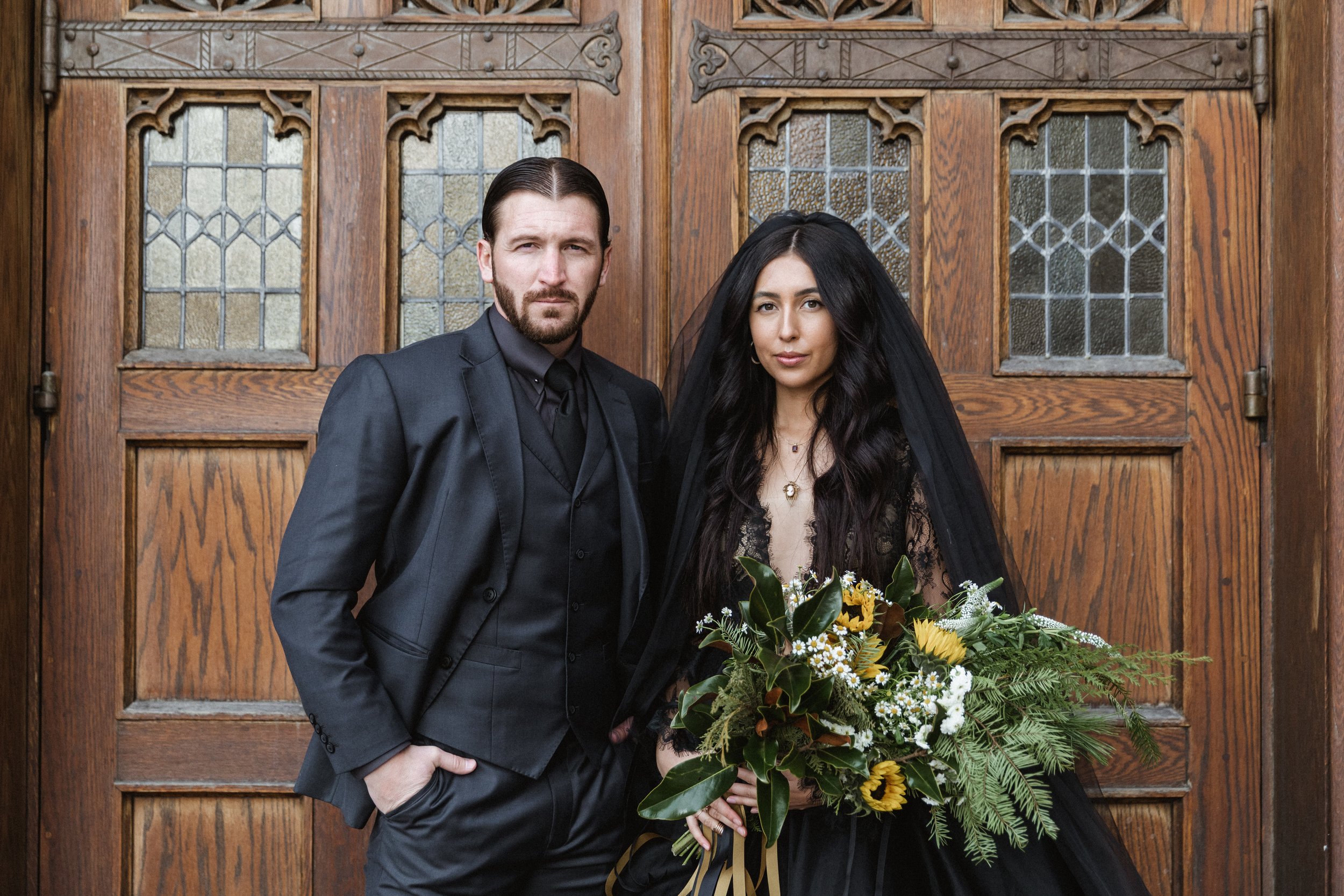 A man and woman stand in front of wooden doors with stained glass windows. The man is dressed in a black suit, and the woman wears a black dress with a veil. The woman holds a large bouquet of yellow, white, and green flowers.