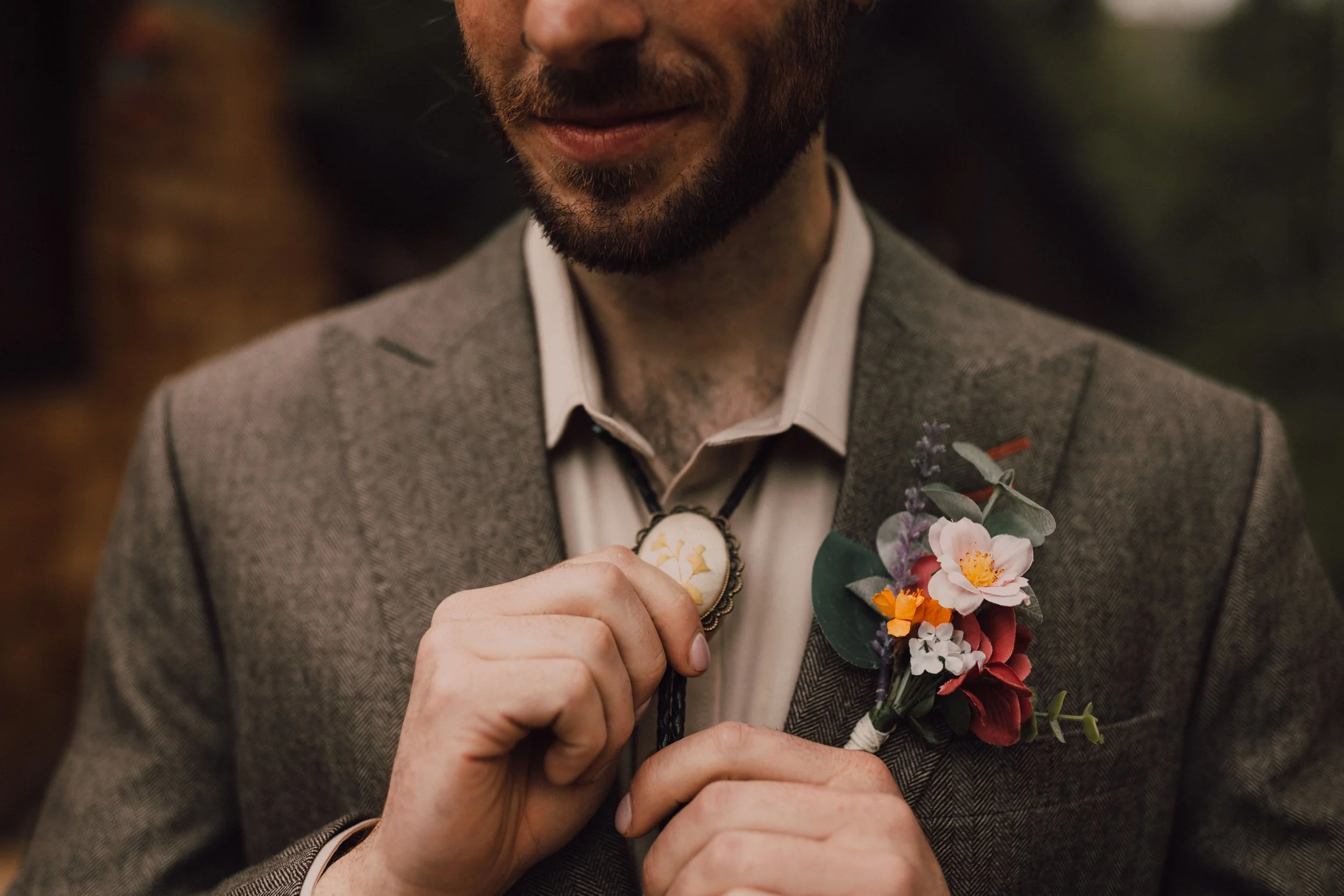 Close-up of a man wearing a brown herringbone jacket with a boutonniere with pink, orange, white flowers and greenery, adjusting a bolo tie.