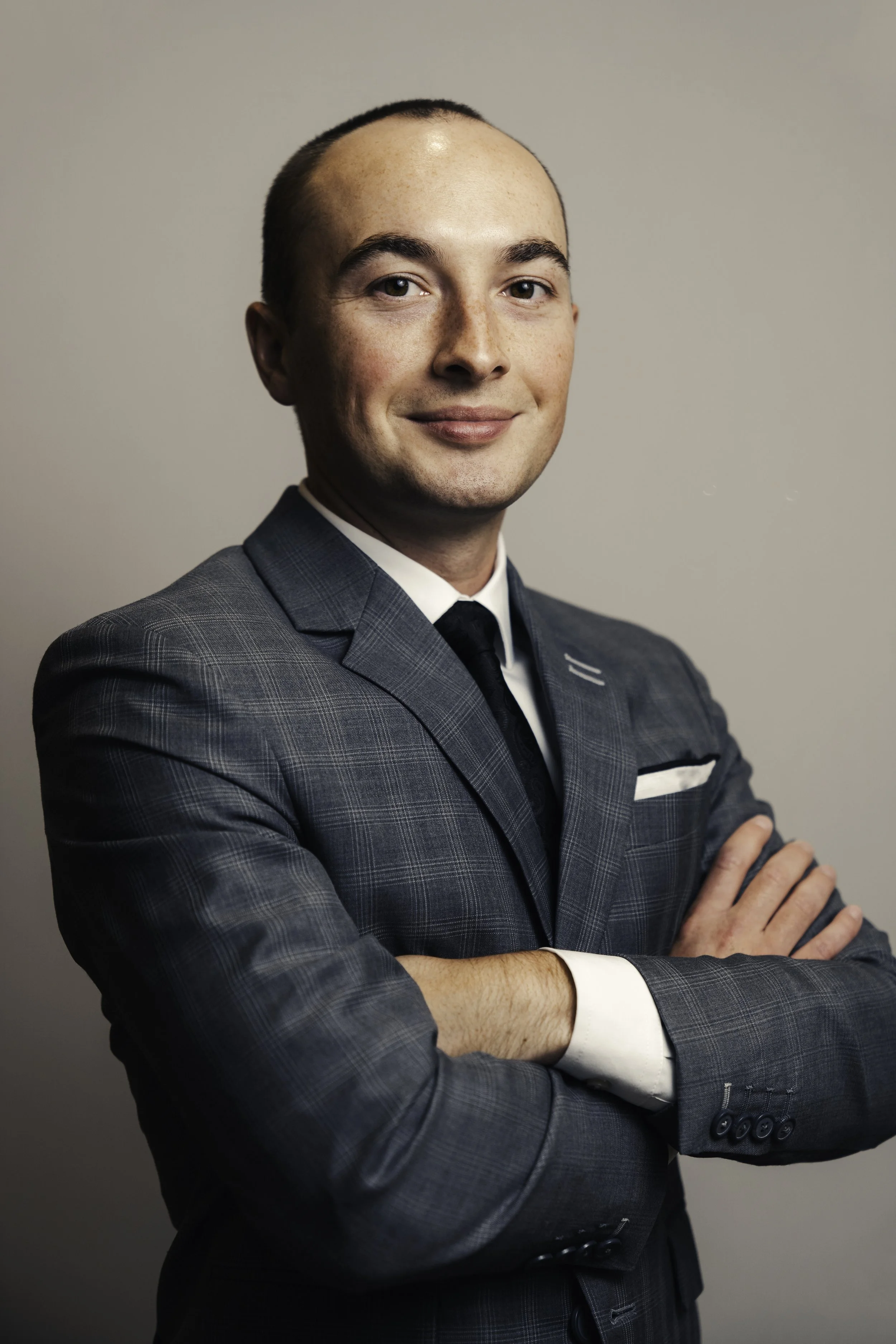 A man in a gray, checkered suit with a white shirt and black tie, standing with his arms crossed, smiling slightly, against a plain light gray background.