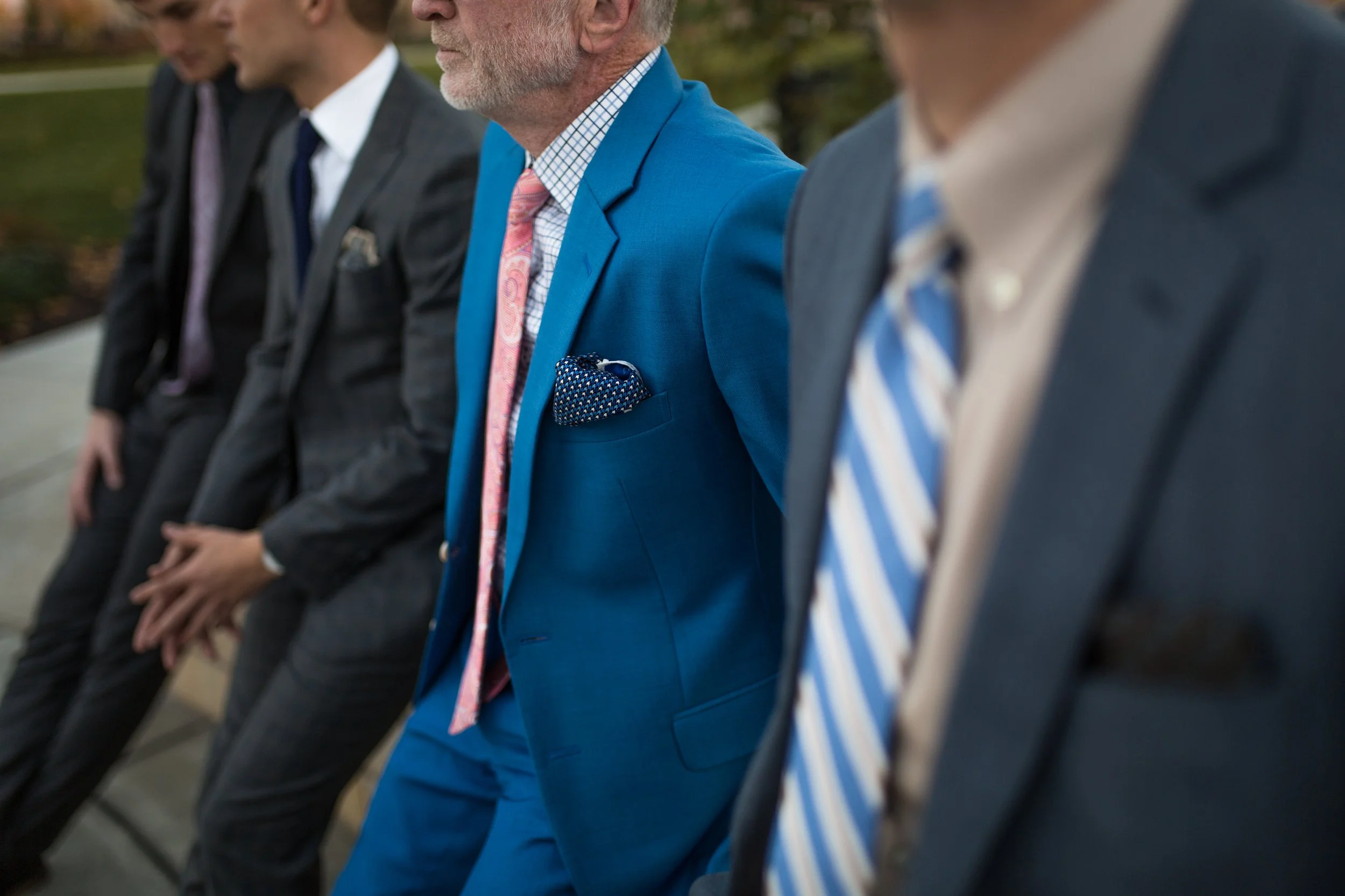 Four men sitting outside wearing suits, with only parts of their faces and torsos visible.