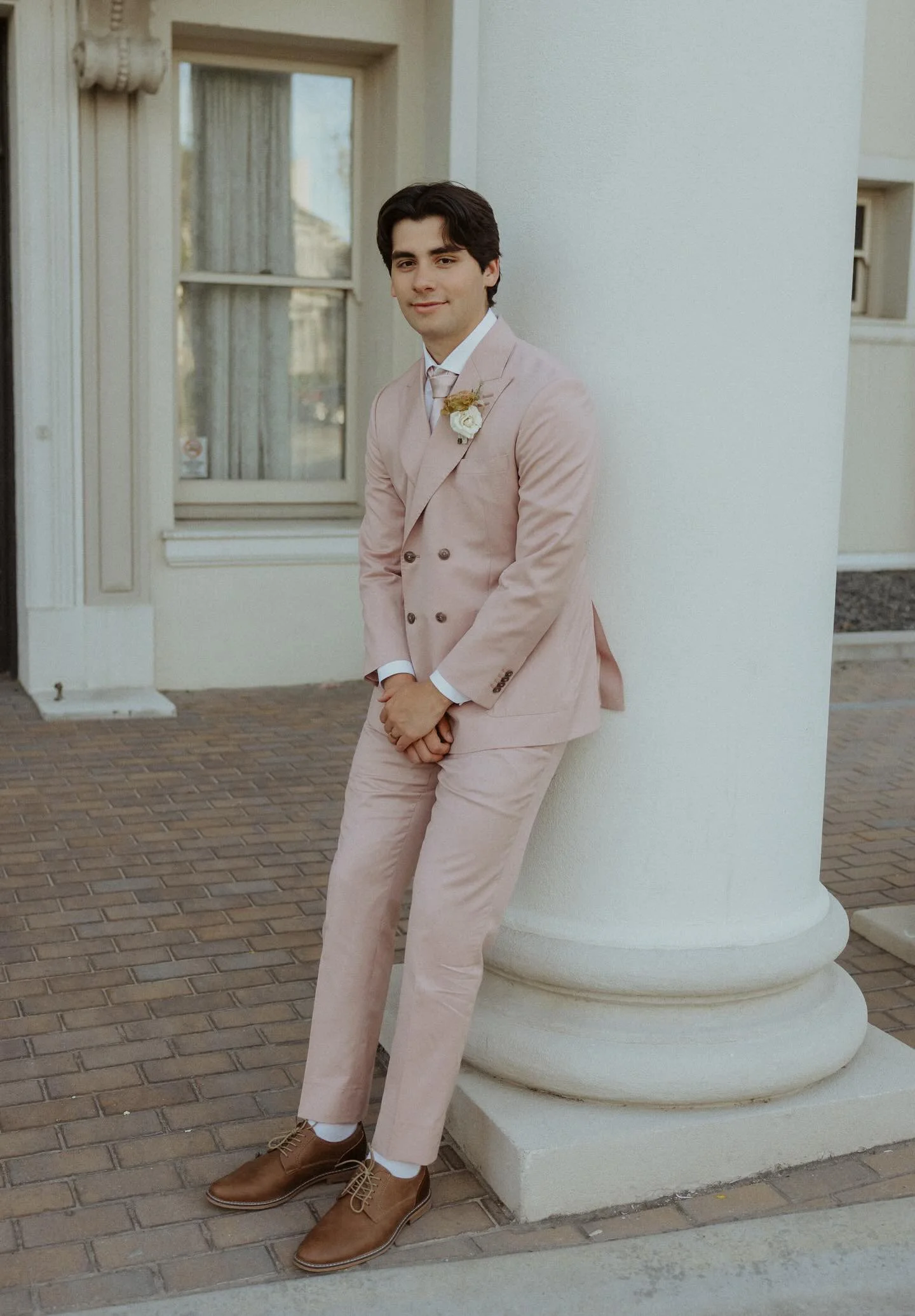 A young man in a pink suit with a boutonniere, leaning against a white pillar outside a building, smiling at the camera.