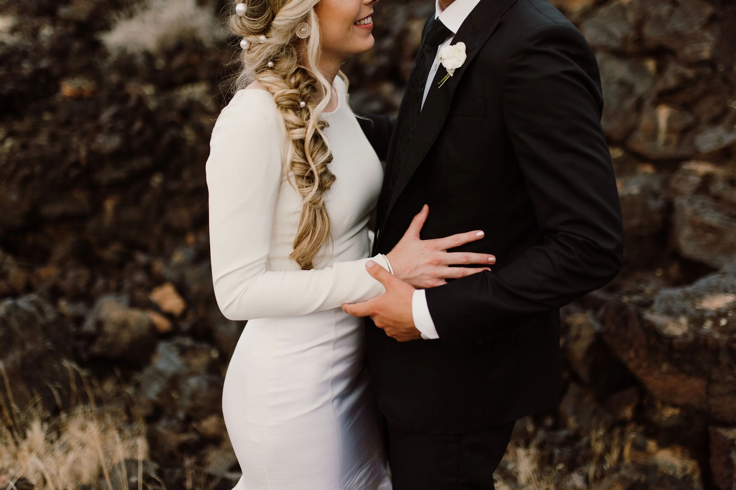 A bride and groom standing close together outdoors, smiling, with their faces near each other, holding hands.