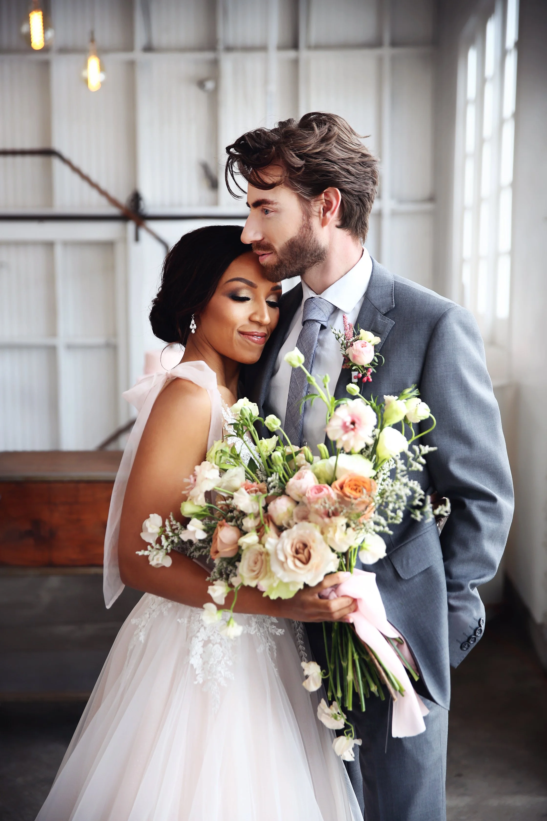 Bride and groom embracing indoors, bride holding a large bouquet of pink, cream, and white flowers, wedding dress with lace details, groom in gray suit and tie, warm smiles, natural light from windows.