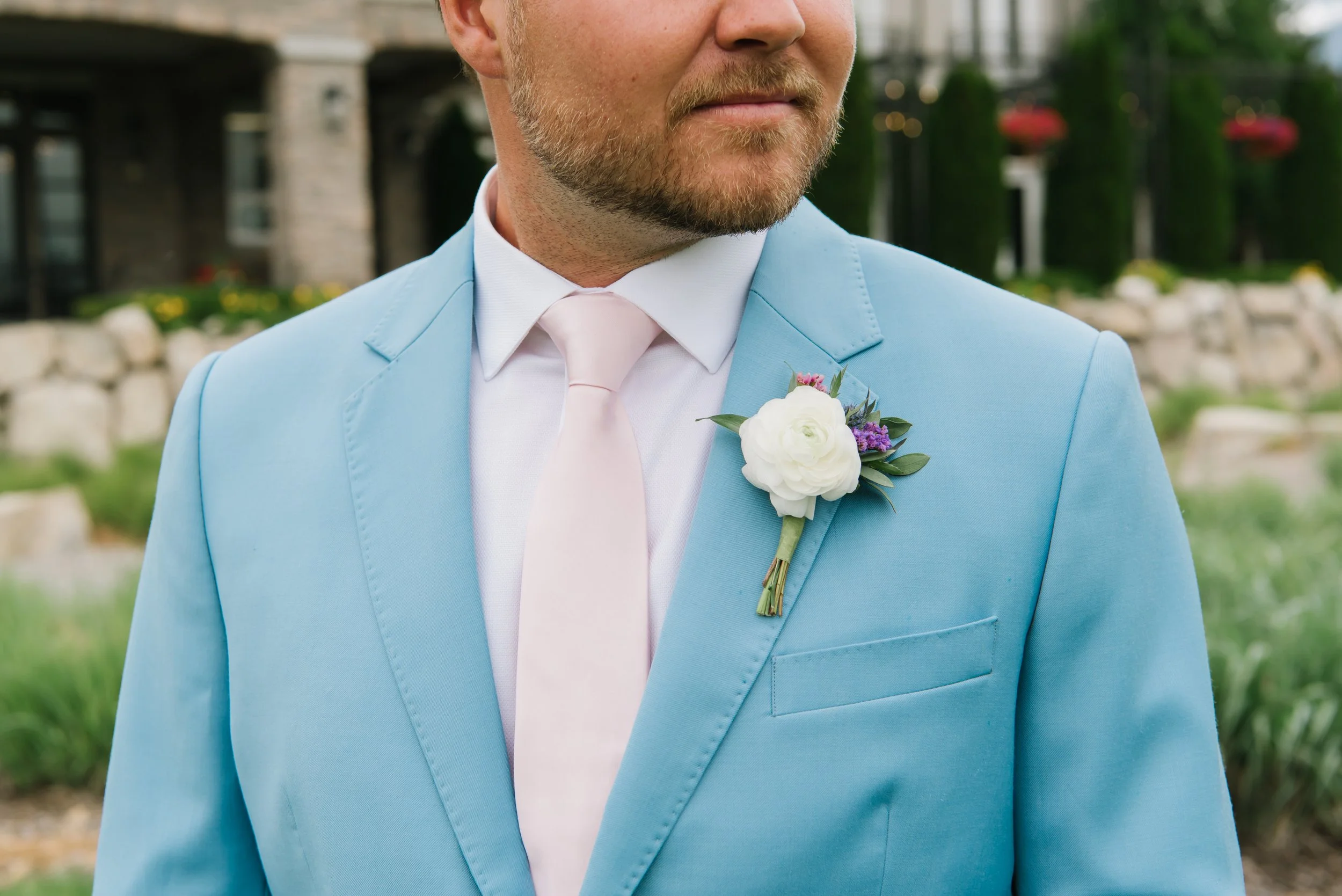 Close-up of a groom wearing a light blue suit with a white dress shirt and a light pink tie, with a white boutonniere pinned to his lapel, outdoors with a blurred background of green grass and trees.