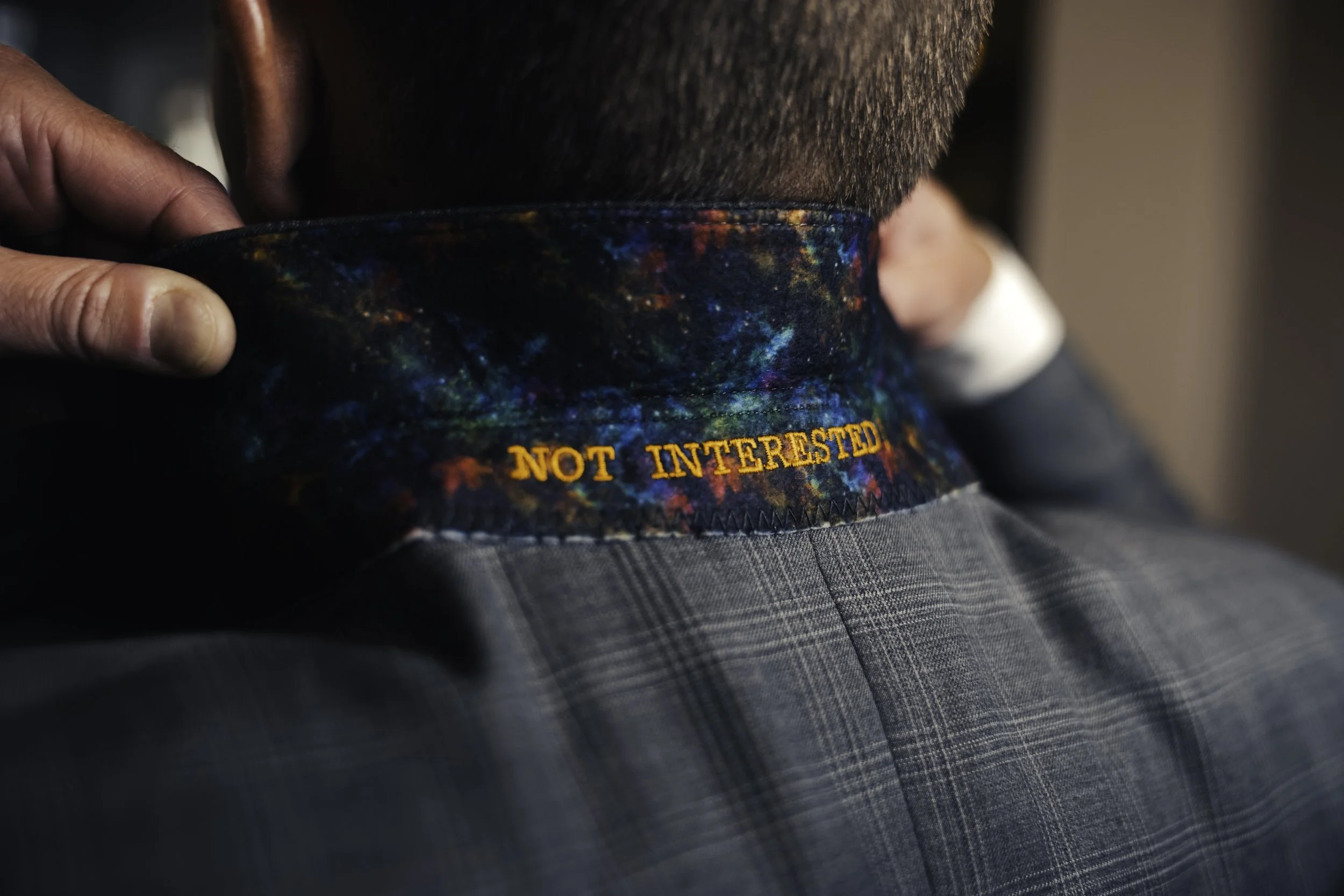 Person adjusting colorful, galaxy-patterned under collar lining with the phrase 'NOT INTERESTED' embroidered in orange at the back of their neck near a grey suit jacket.