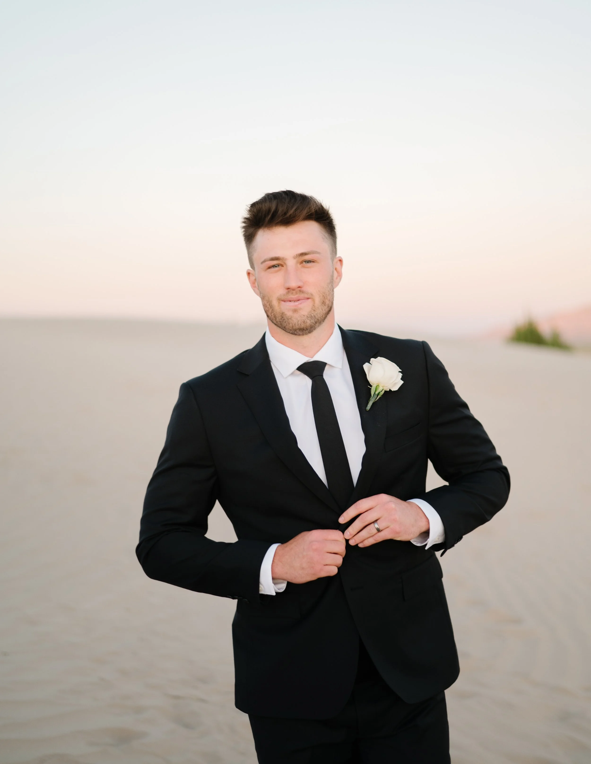 A man in a black tuxedo with a white dress shirt and black tie, standing on a beach during sunset, smiling at the camera.