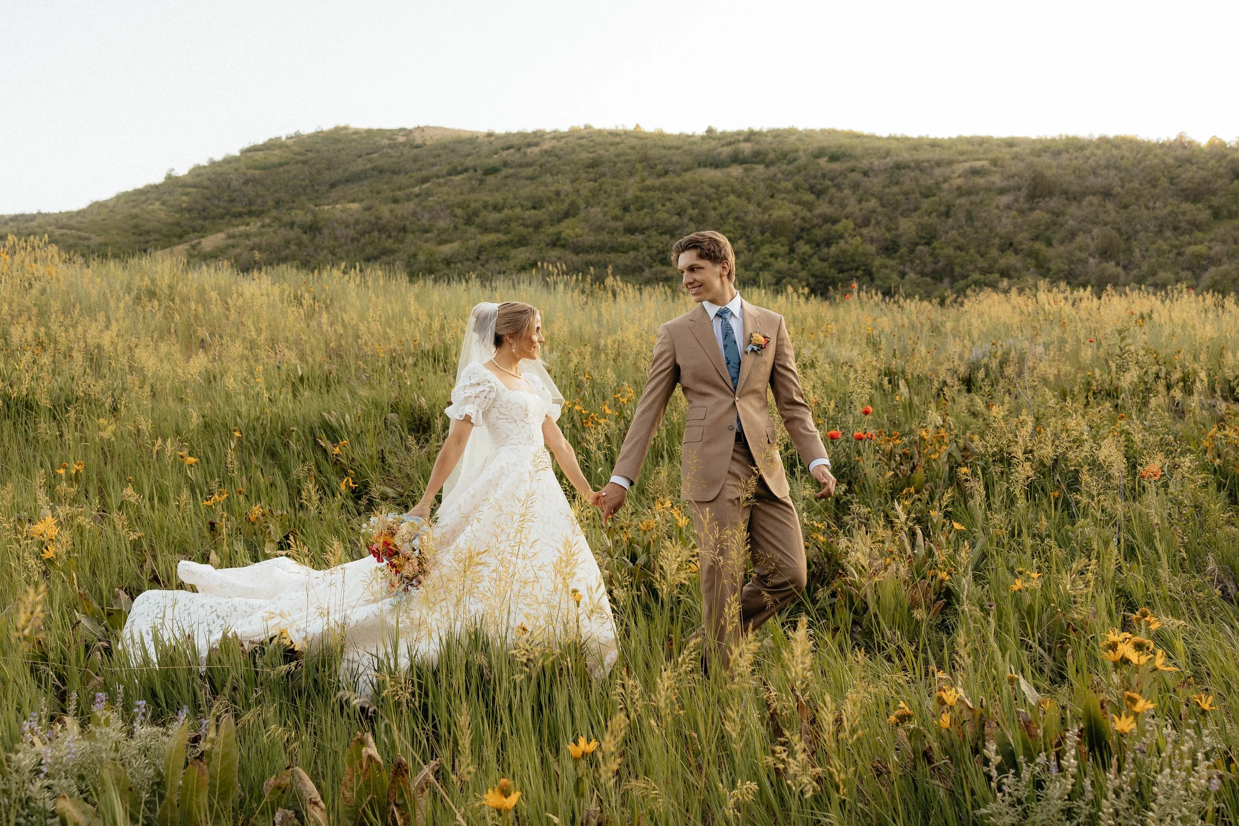 A bride and groom holding hands and walking through a field of yellow flowers with hills and a clear sky in the background.