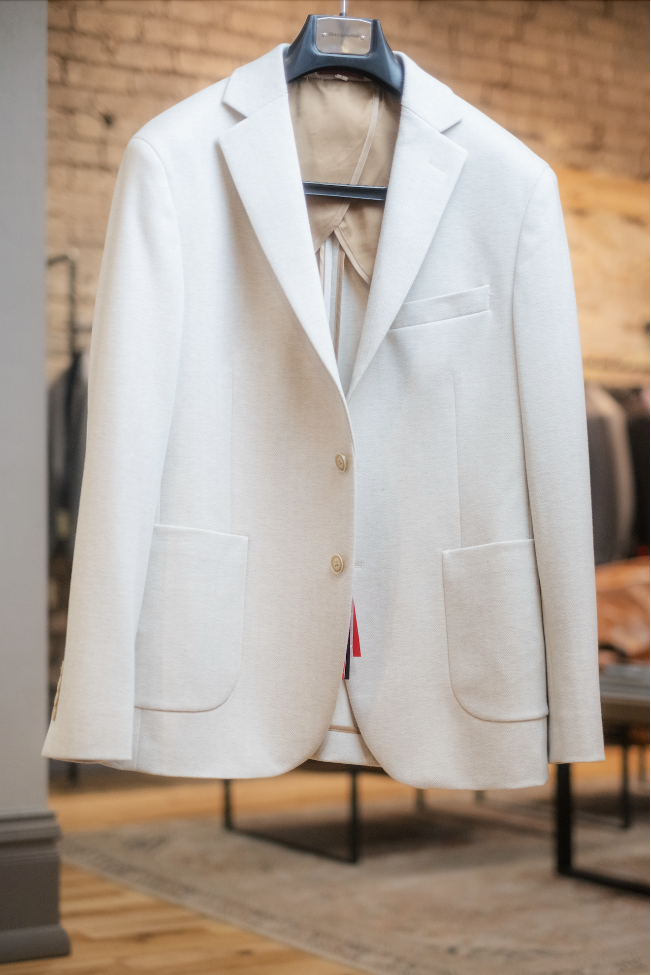 A white sport coat hanging on a black hanger inside a clothing store, with a brick wall in the background.
