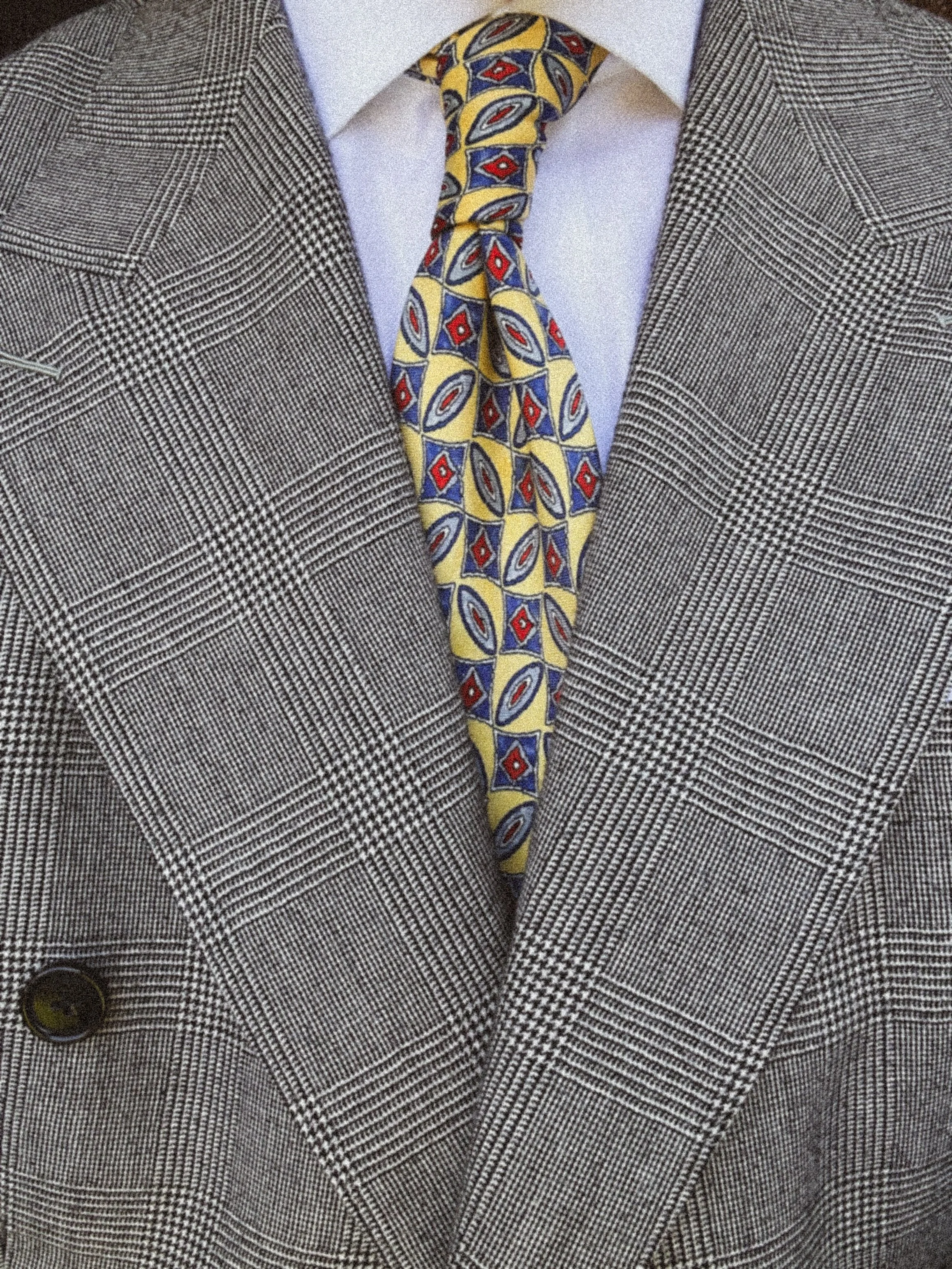 Close-up of a man wearing a grey glen plaid suit jacket, a white dress shirt, a colorful patterned tie, and a dark button on his jacket.