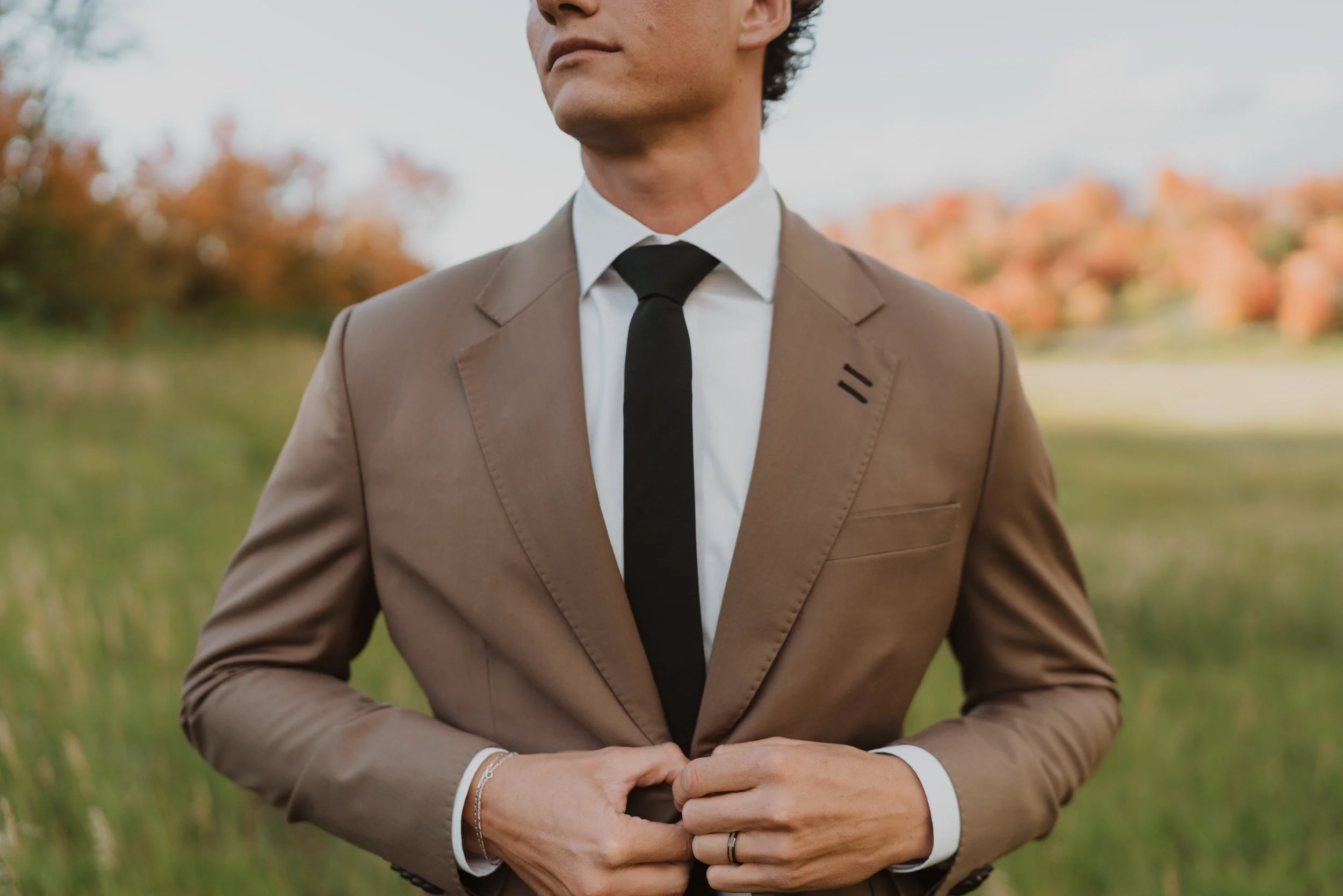 A person dressed in a brown suit with a white shirt and black tie, standing outdoors with autumn foliage in the background.