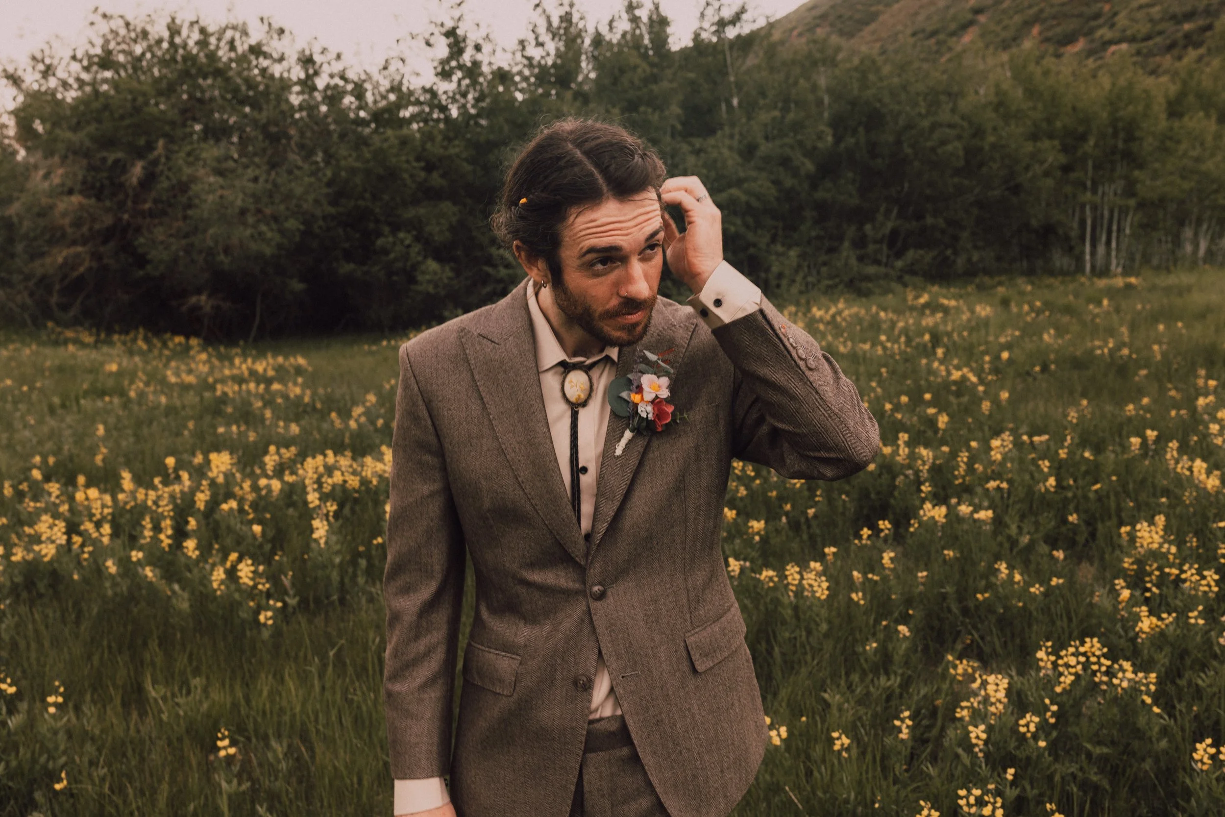 A man in a brown suit with a floral boutonniere stands in a field of yellow flowers with trees and hills in the background, adjusting his hair.