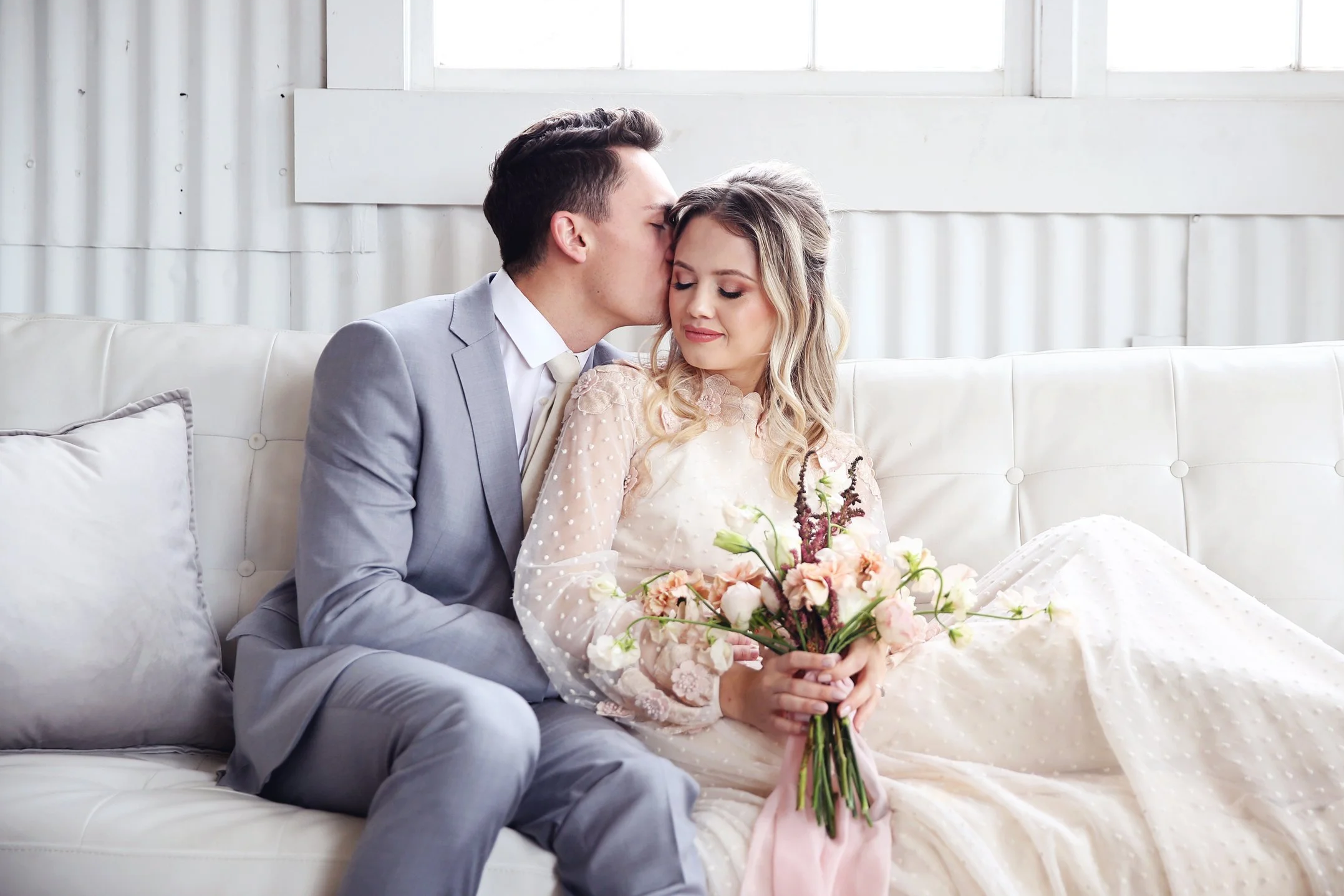 A bride and groom sitting on a white sofa, with the groom kissing the bride on her forehead. The bride is holding a bouquet of pink and white flowers, and they are both dressed in wedding attire. The background features white walls and a large window