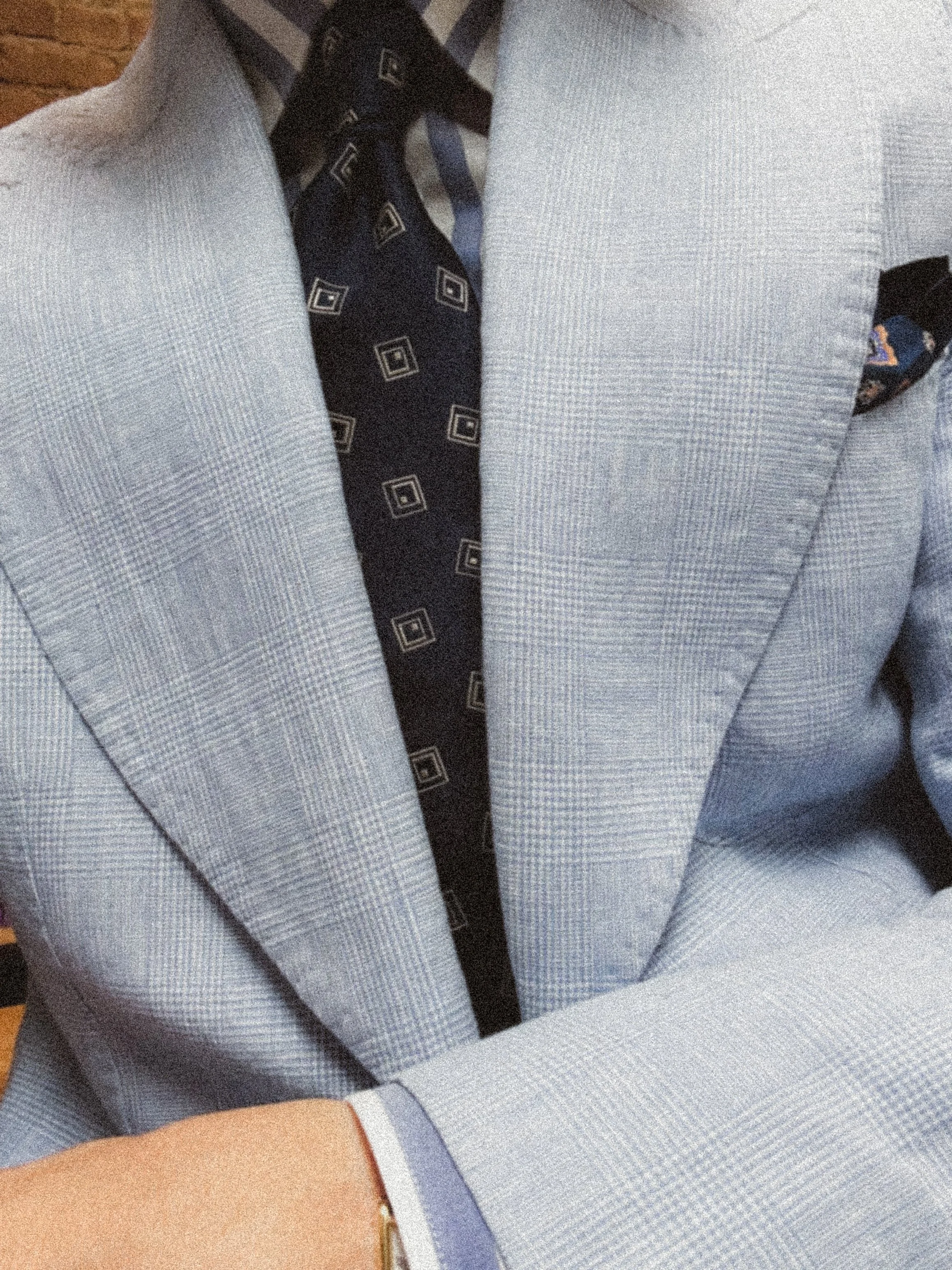 A person wearing a light gray checkered suit and a black tie with a geometric pattern. They are also wearing a watch and a ring on their left hand.