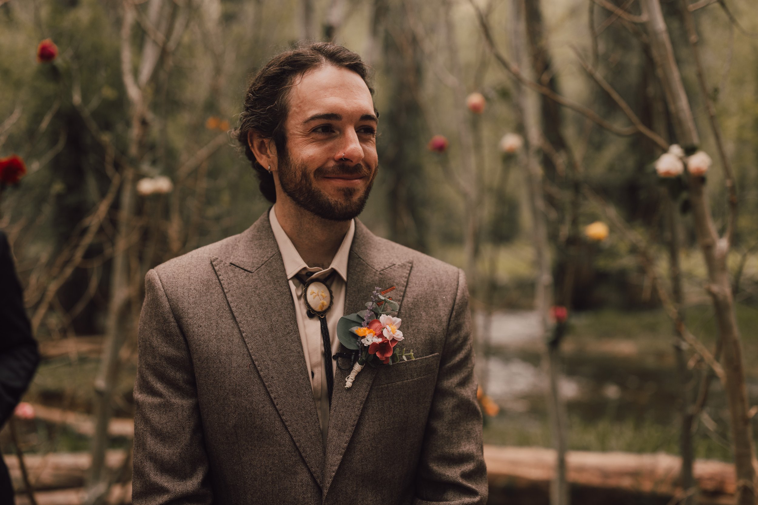 A man with a beard and long hair wearing a brown suit with a floral boutonniere, smiling outdoors in a garden with hanging flowers.
