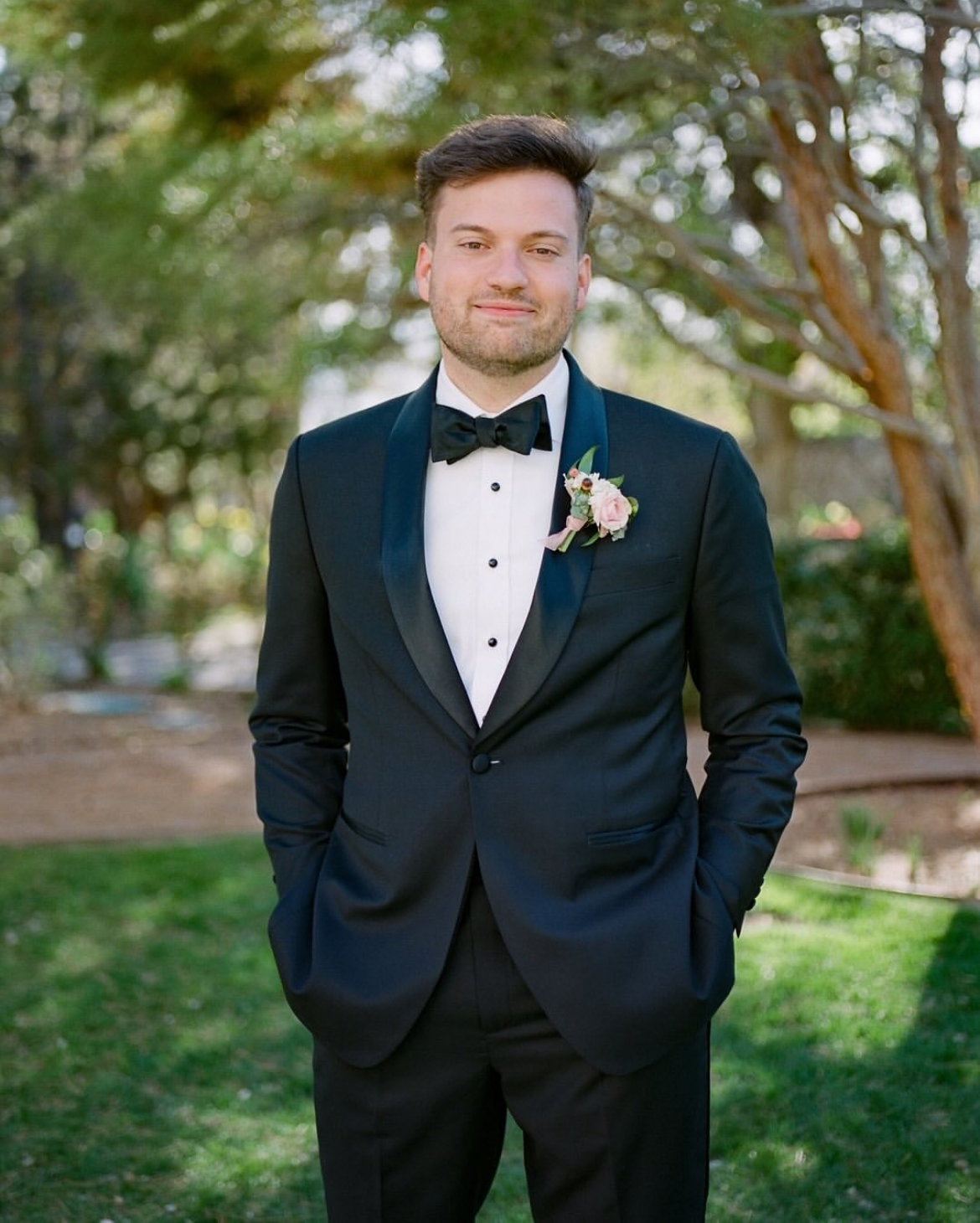 A man in a black tuxedo with a bow tie and a boutonniere, standing outdoors in a garden, smiling at the camera.