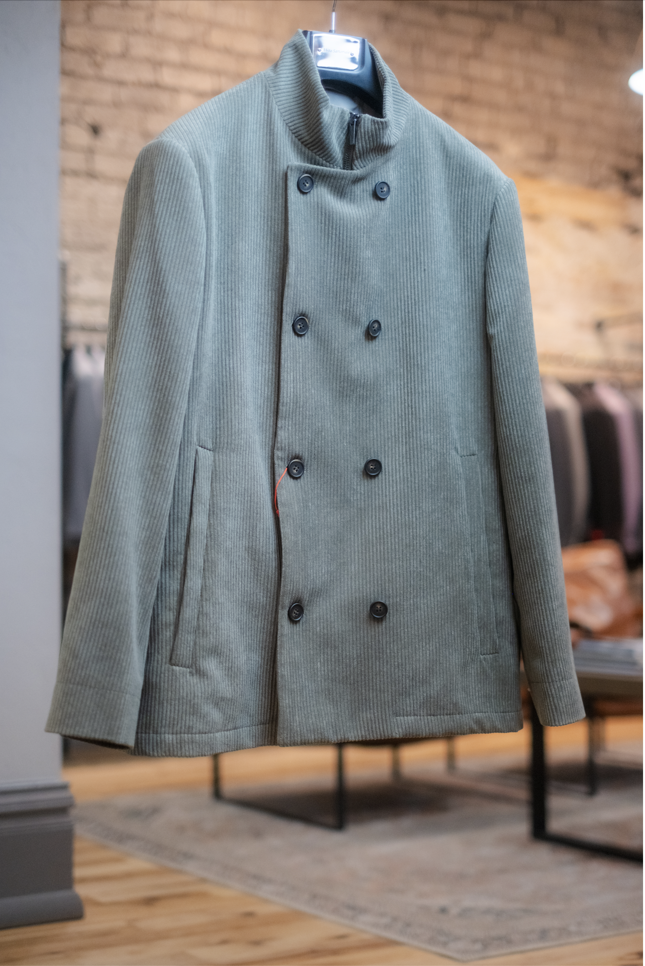 A gray, double-breasted, high-collared corduroy jacket hanging on a black hanger in a clothing store, with a brick wall background and wooden floor.