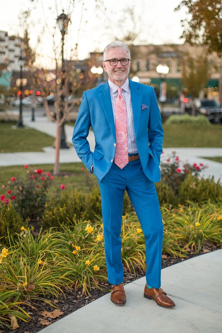 A man in a bright blue suit with a pink paisley tie, standing outdoors in a park or garden area during daytime, smiling with hands in pockets.