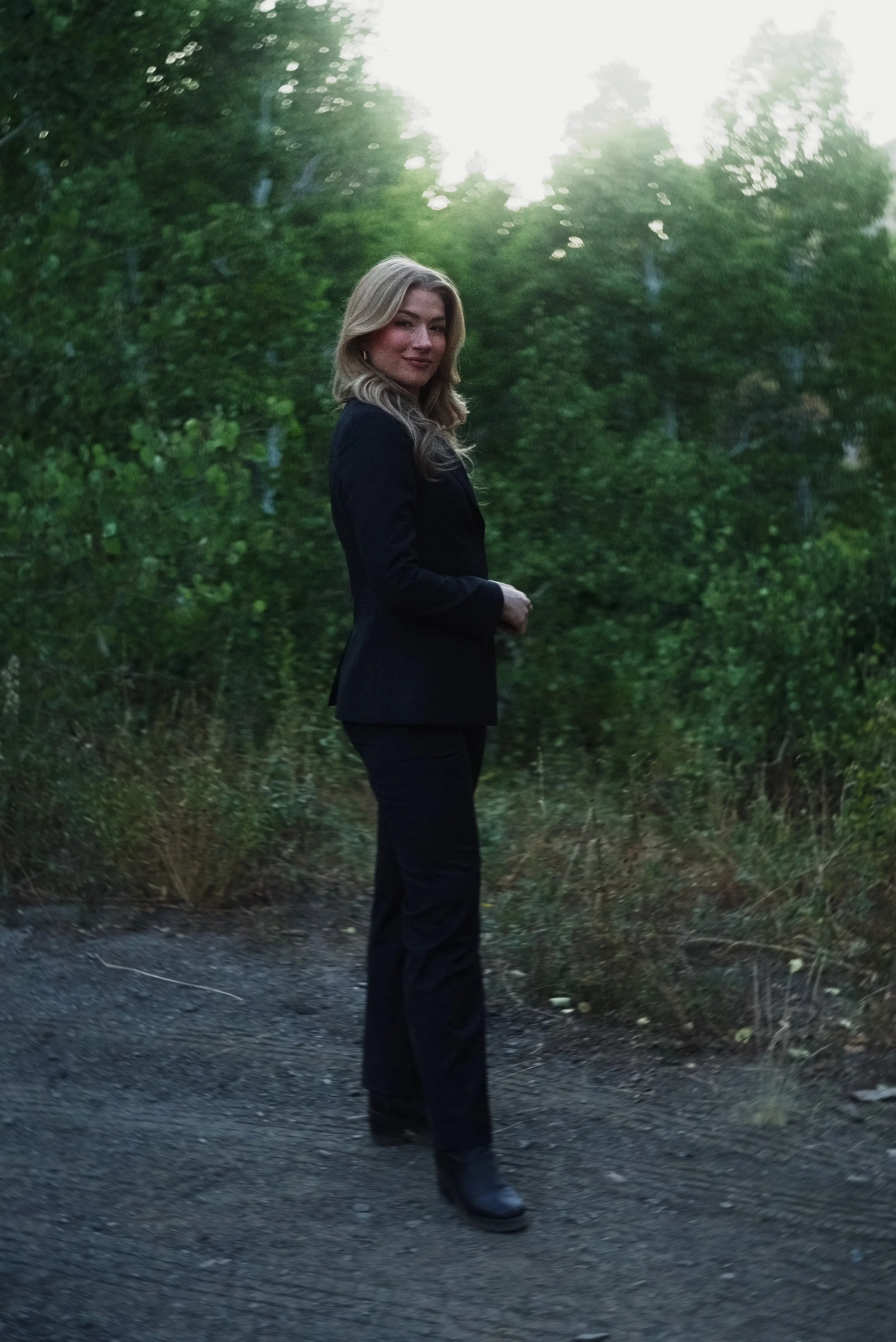 A woman with blonde hair wearing a black blazer and pants stands outdoors on a dirt path surrounded by green trees, with sunlight filtering through the leaves in the background.